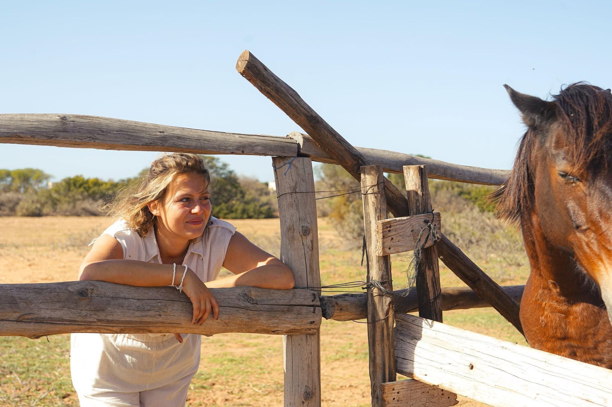 A woman with light skin and blonde hair gazes at a brown horse behind a wooden fence at an outdoor farm setting on a sunny day.