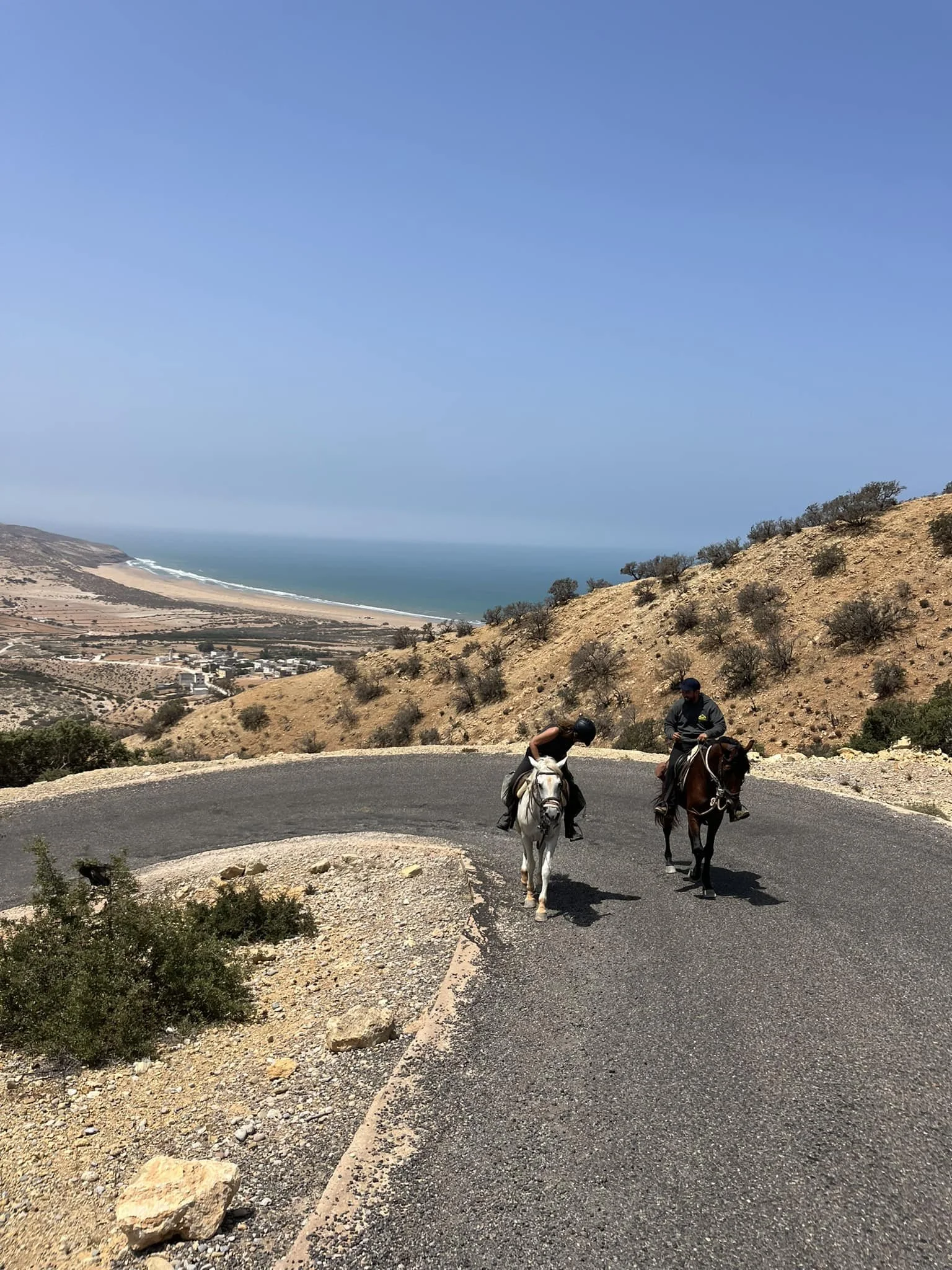 Two people riding horses on a mountain road overlooking a coastal landscape with a sandy beach and the ocean in the background. The sky is clear with no clouds, and the terrain is arid with sparse vegetation.