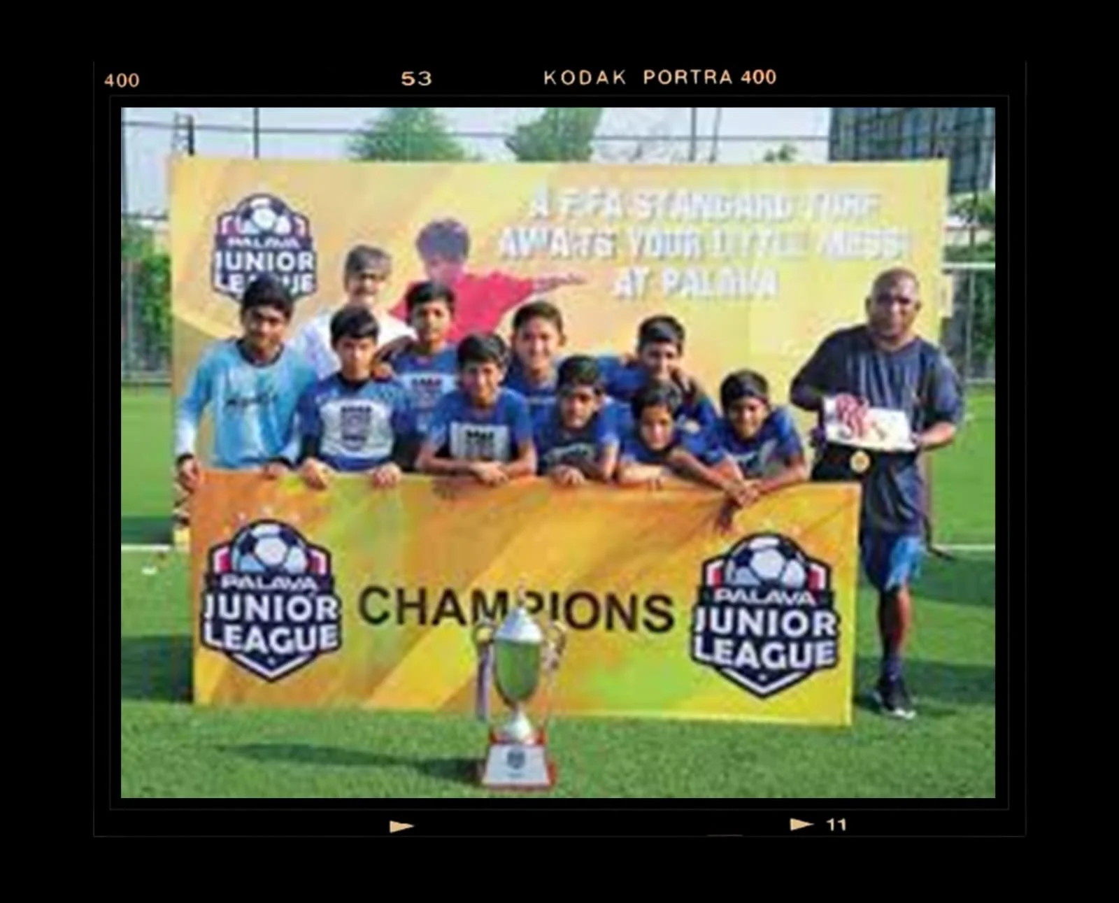 A youth soccer team posing with a coach, holding a banner that says "Champions" with a trophy and a cake in front of them, celebrating their win in the Saglina Junior League.