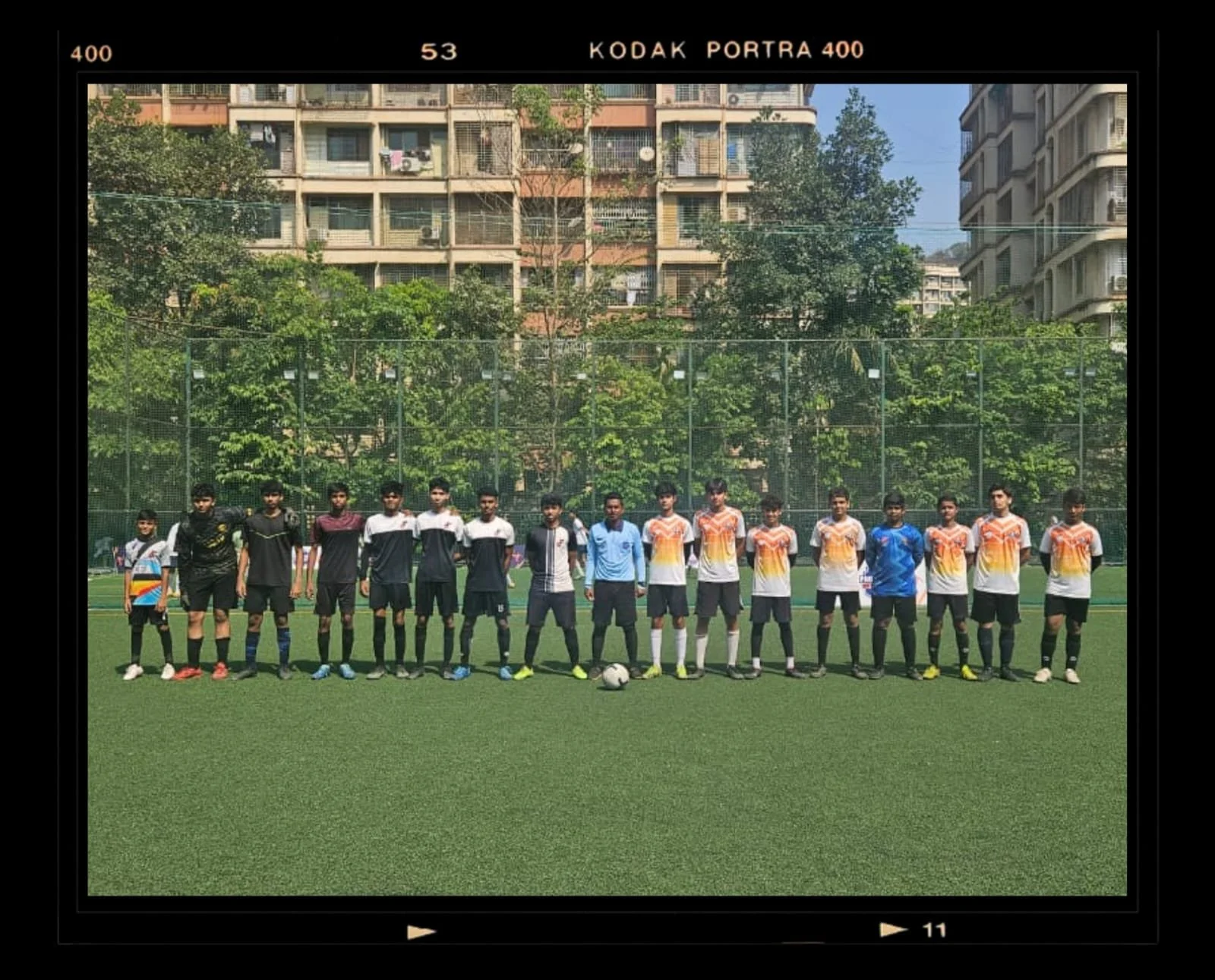 Team of boys standing in a line on a green football field, with buildings and trees in the background.