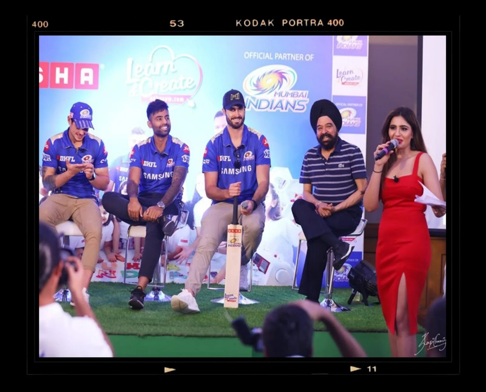 A group of five people sitting and standing on a stage during a promotional event, with a backdrop featuring the Mumbai Indians cricket team logo and sponsors, some wearing Mumbai Indians jerseys and a woman in a red dress speaking into a microphone.