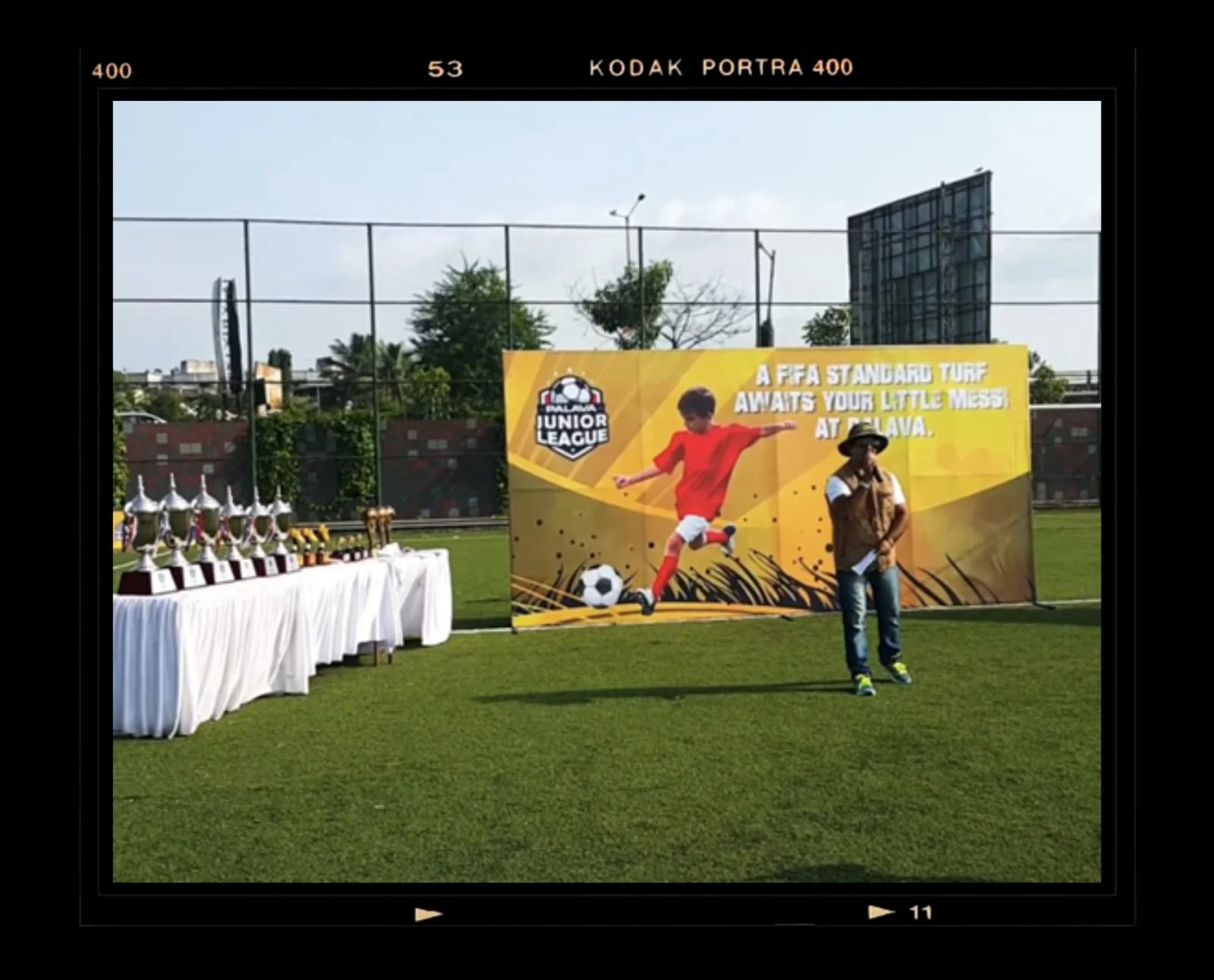 An outdoor sports field with trophies on a table to the left, and a large colorful banner in the center displaying a young boy playing soccer with the text "A FFA STANDARD TURF AWAITS YOUR LITTLE MESS!" On the right, a person wearing a hat, sunglasses, and casual clothing is standing in front of the banner. There is a black fence, trees, and buildings in the background.