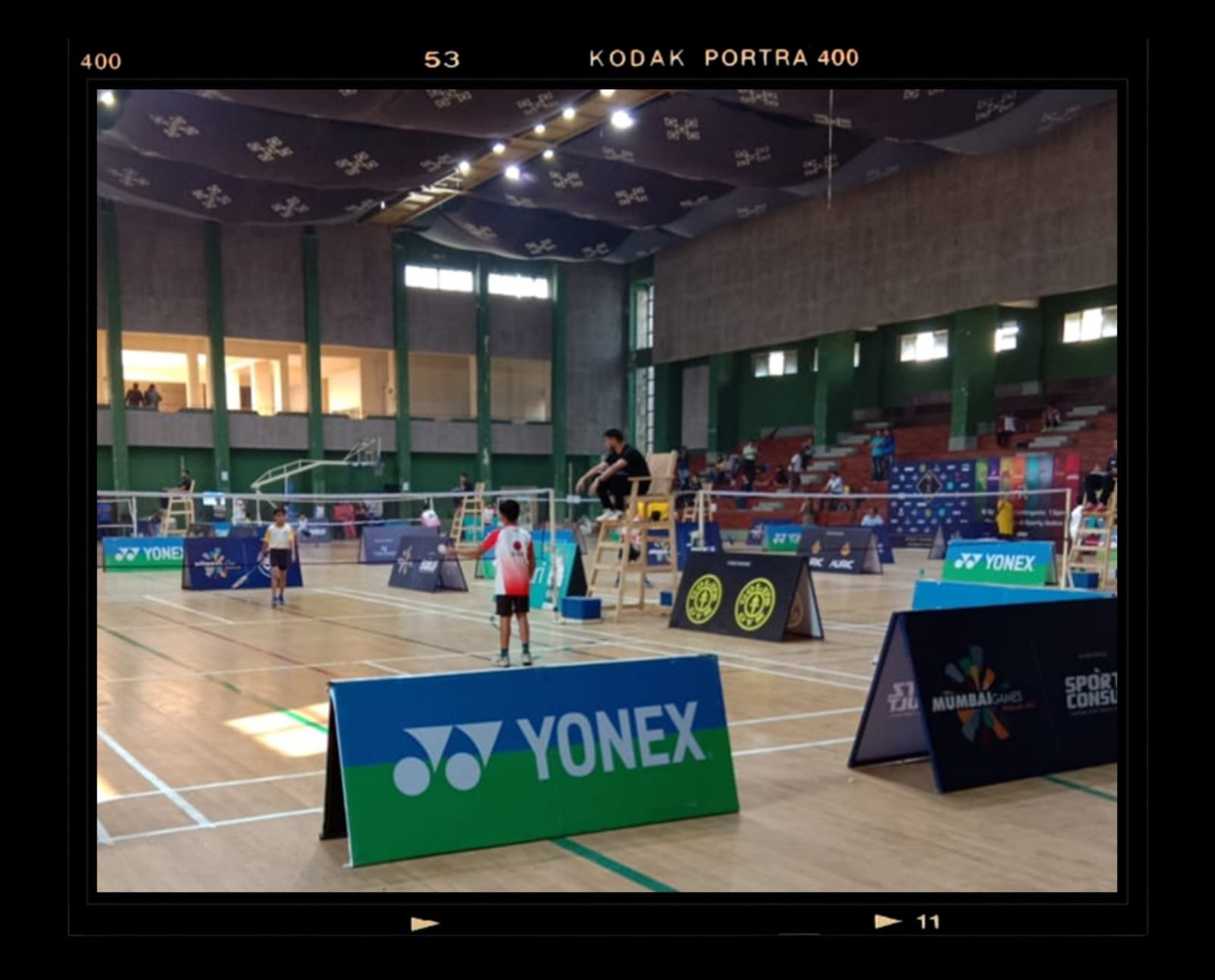 Indoor badminton court with players and umpire. Banners and advertisements for Yonex and other sponsors are visible, and some spectators are seated in the background.
