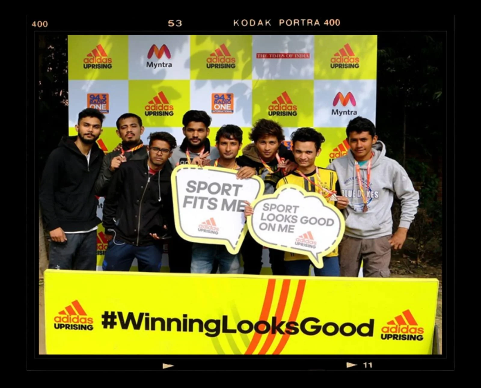 Group of young men standing outdoors, holding signs that say "Sport Fits Me" and "Sport Looks Good On Me", in front of a yellow backdrop with the hashtag "WinningLooksGood" and logos of Adidas Uprising, Myntra, and Radio One, with trees visible in the background.