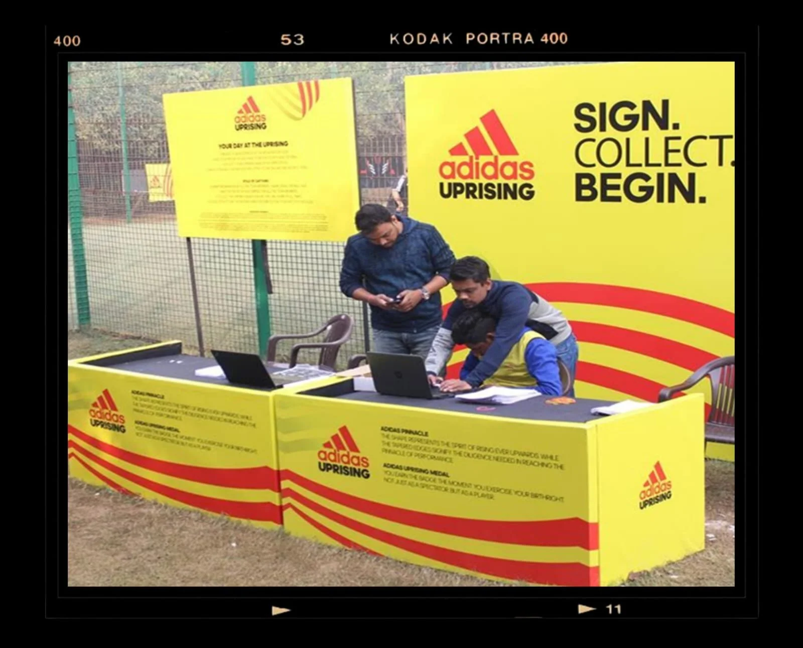 Three young men are gathered around a yellow table with red stripes and Adidas Uprising branding. Two are working on laptops, while the third looks at his phone. Behind them, a large yellow Adidas Uprising sign with bold black text reads "SIGN. COLLECT. BEGIN." A yellow poster with similar branding is on a fence in the background, and there are additional chairs and a netted sports field behind them.