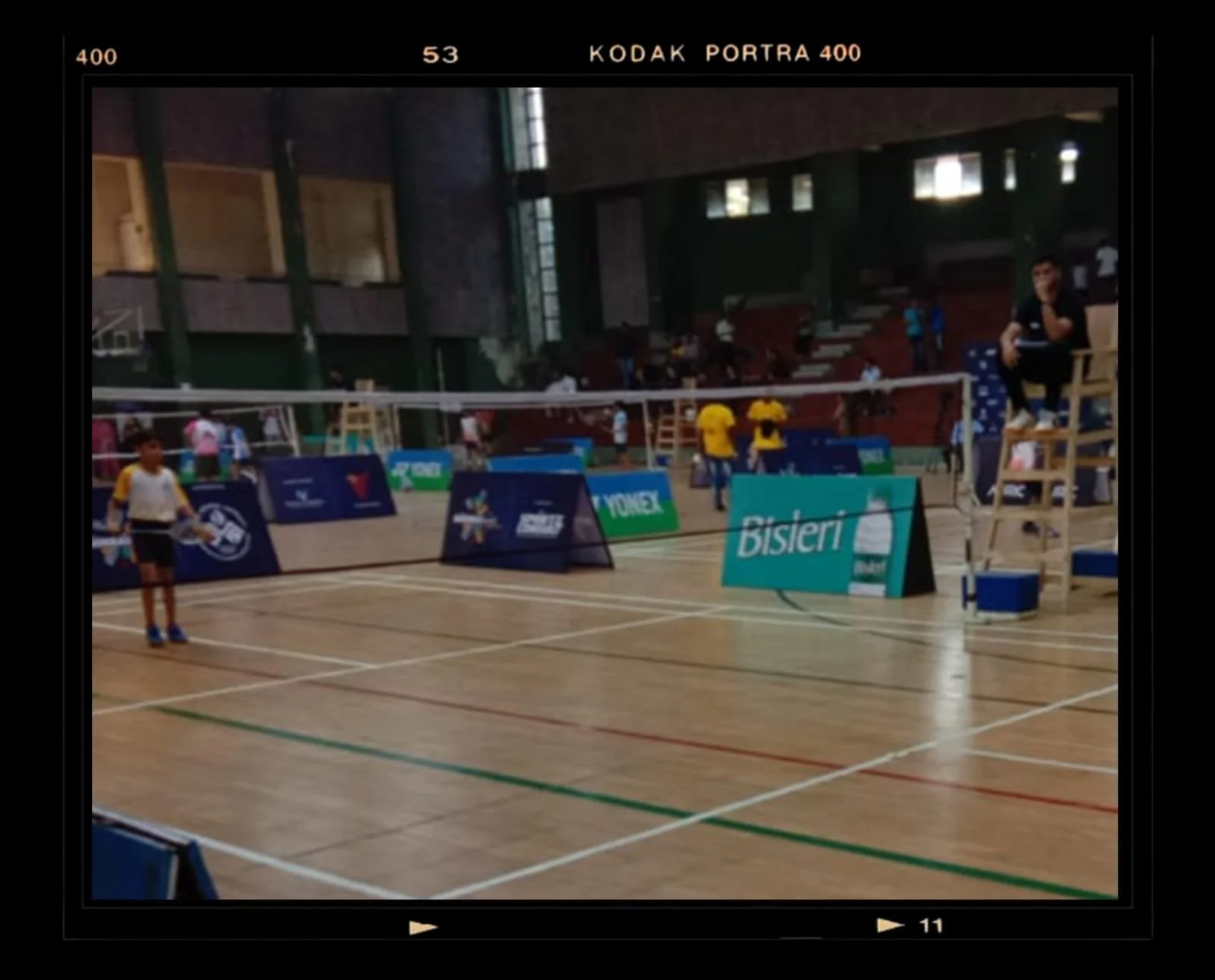 Indoor badminton court with players, referee chair, and banners, under a high ceiling with windows, during a match or practice session.