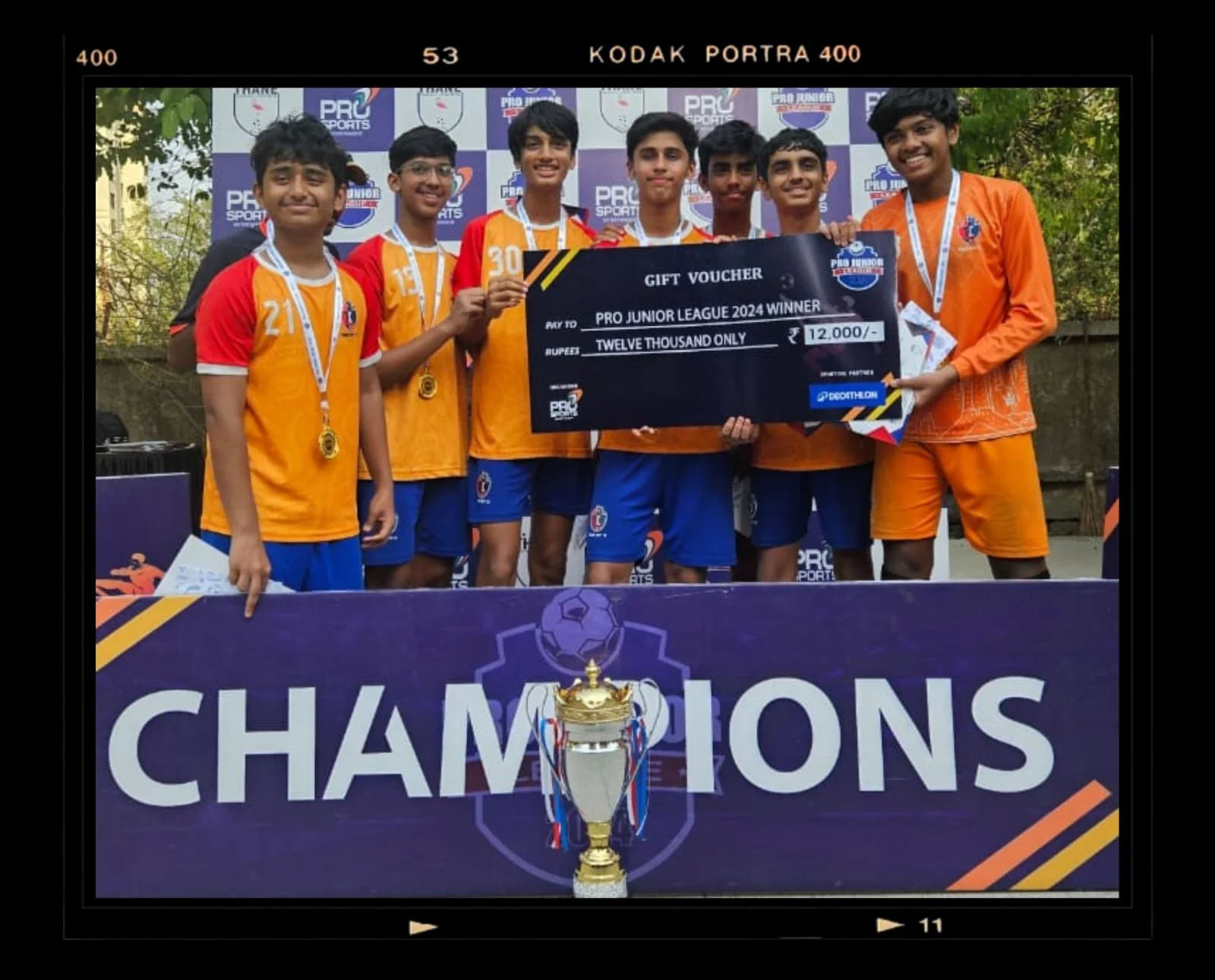 Group of young boys in sports uniforms celebrating their victory at a soccer championship, holding a large gift voucher for 12,000 rupees, with medals around their necks, standing behind a purple banner that reads 'Champions' with a trophy in front of it.