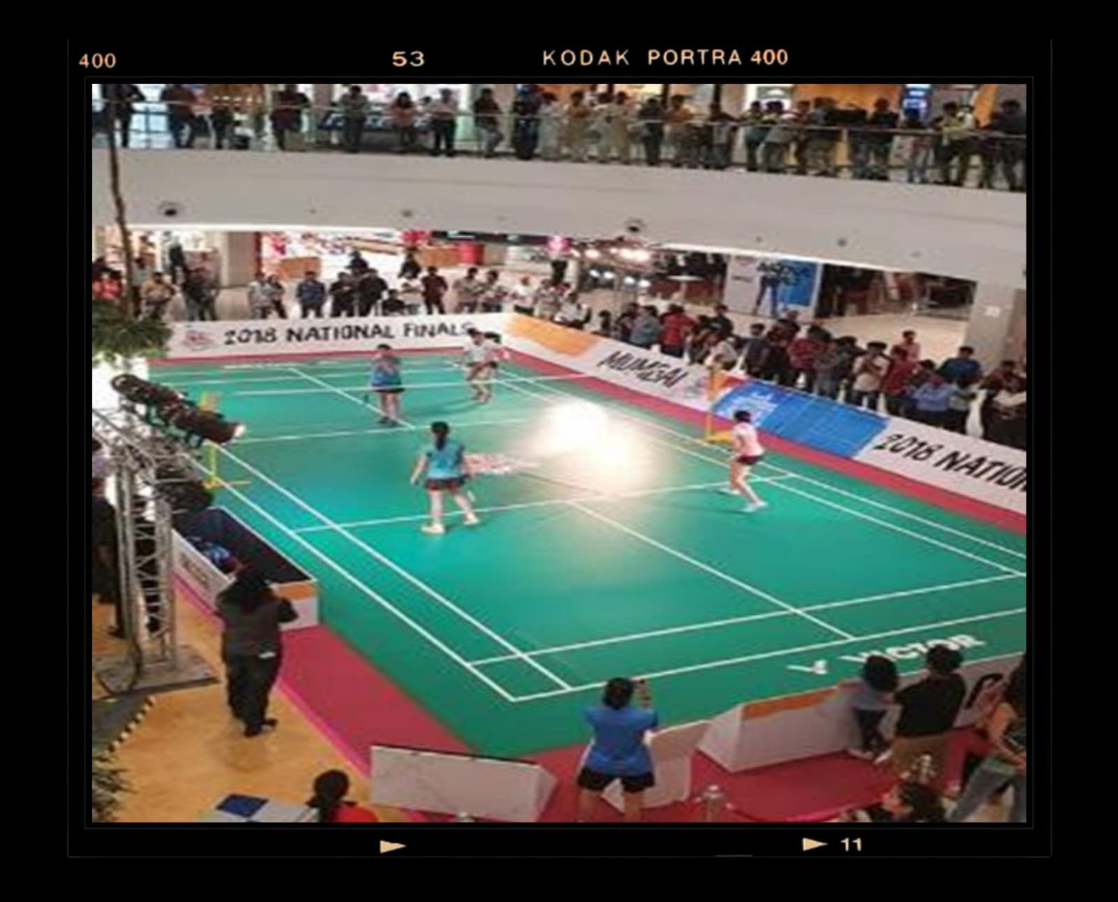 Indoor badminton court with players in action and spectators watching from the sidelines. Banners around the court display '2018 NATIONAL FINAL'.