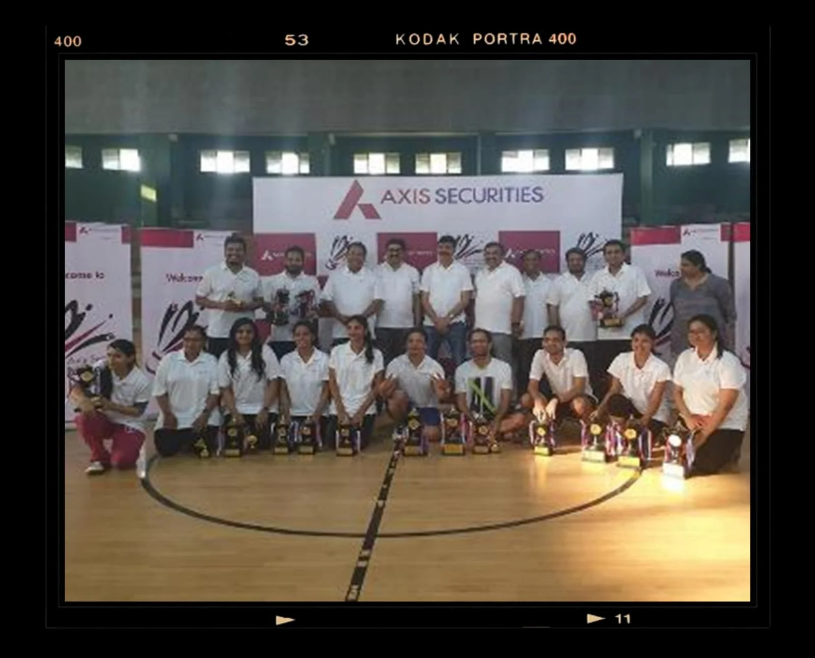 A group of people posing for a photo in an indoor gymnasium, with trophies in front of them, and a banner displaying 'AXIS SECURITIES' behind them.