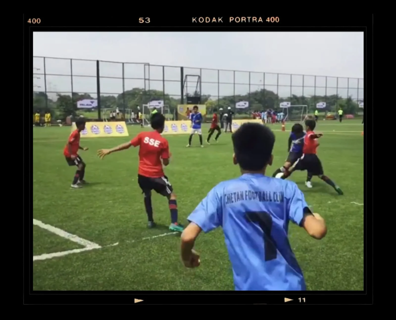 Young kids playing soccer on a field during daytime, with one child in a light blue jersey in the foreground, others in red and blue jerseys, spectators and goalposts in the background.