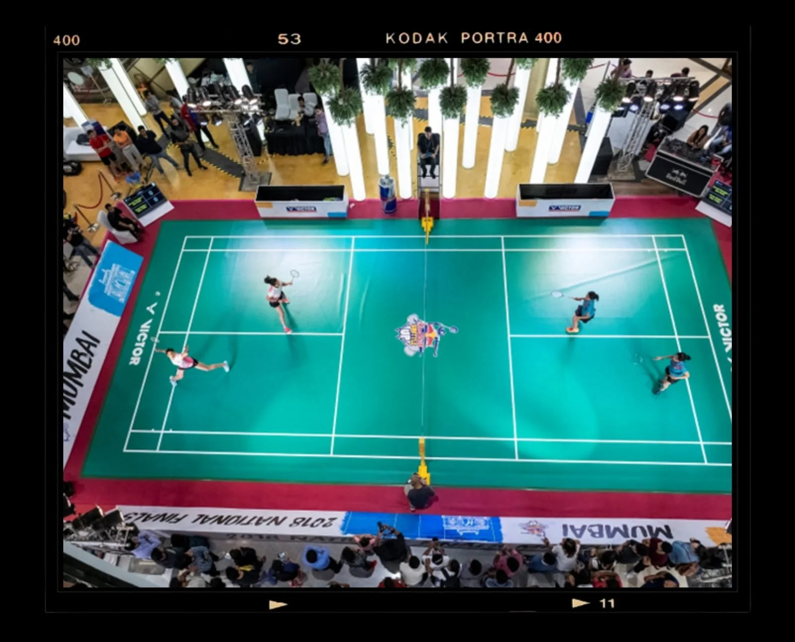 A badminton match taking place on a court with players actively engaged, with spectators and event staff around.