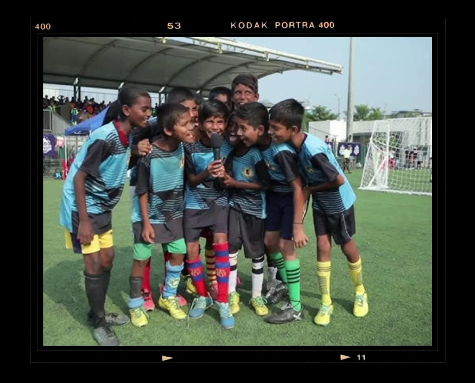 A group of young children in sports uniforms gathered on a soccer field, celebrating with a man holding a microphone. The children are smiling and hugging each other, with soccer goals and a large crowd in the background.