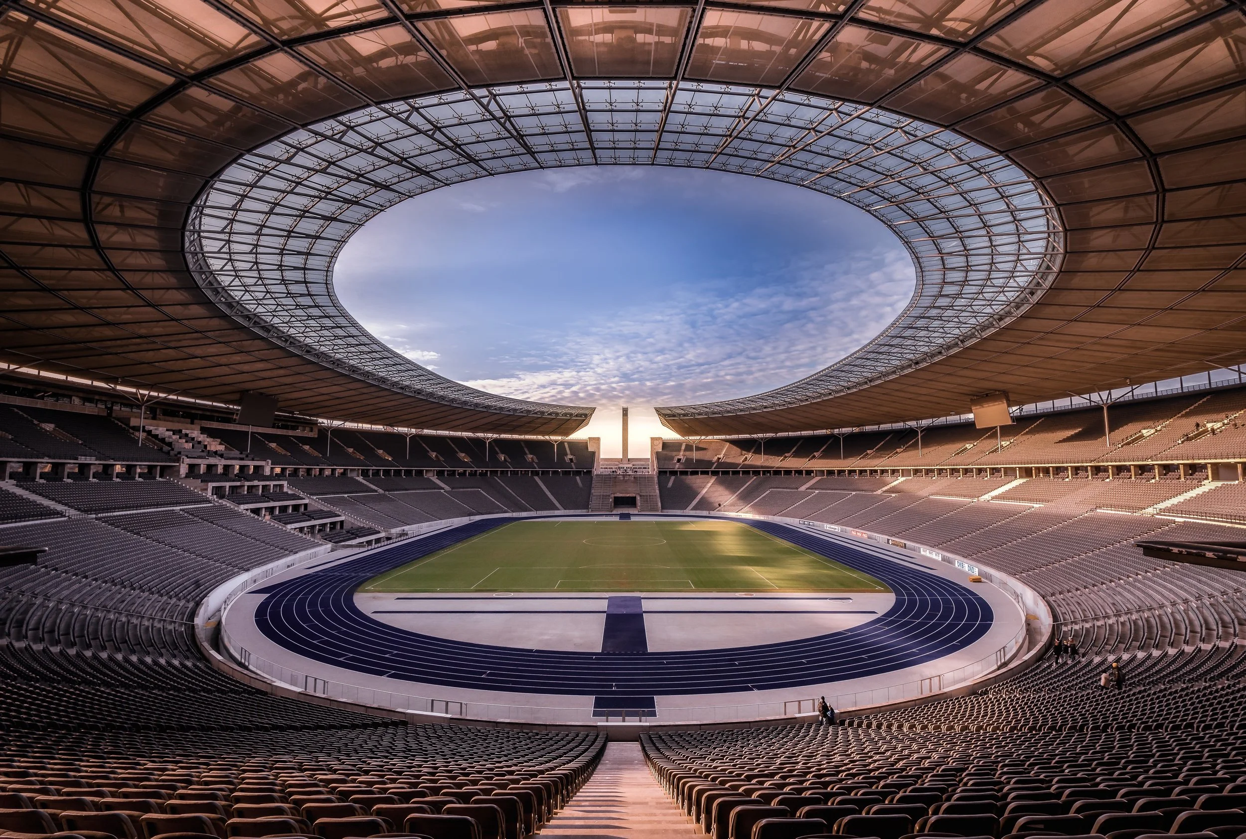 Empty modern stadium with a green soccer field and a track, viewed from the stands with a circular roof structure and cloudy sky overhead.