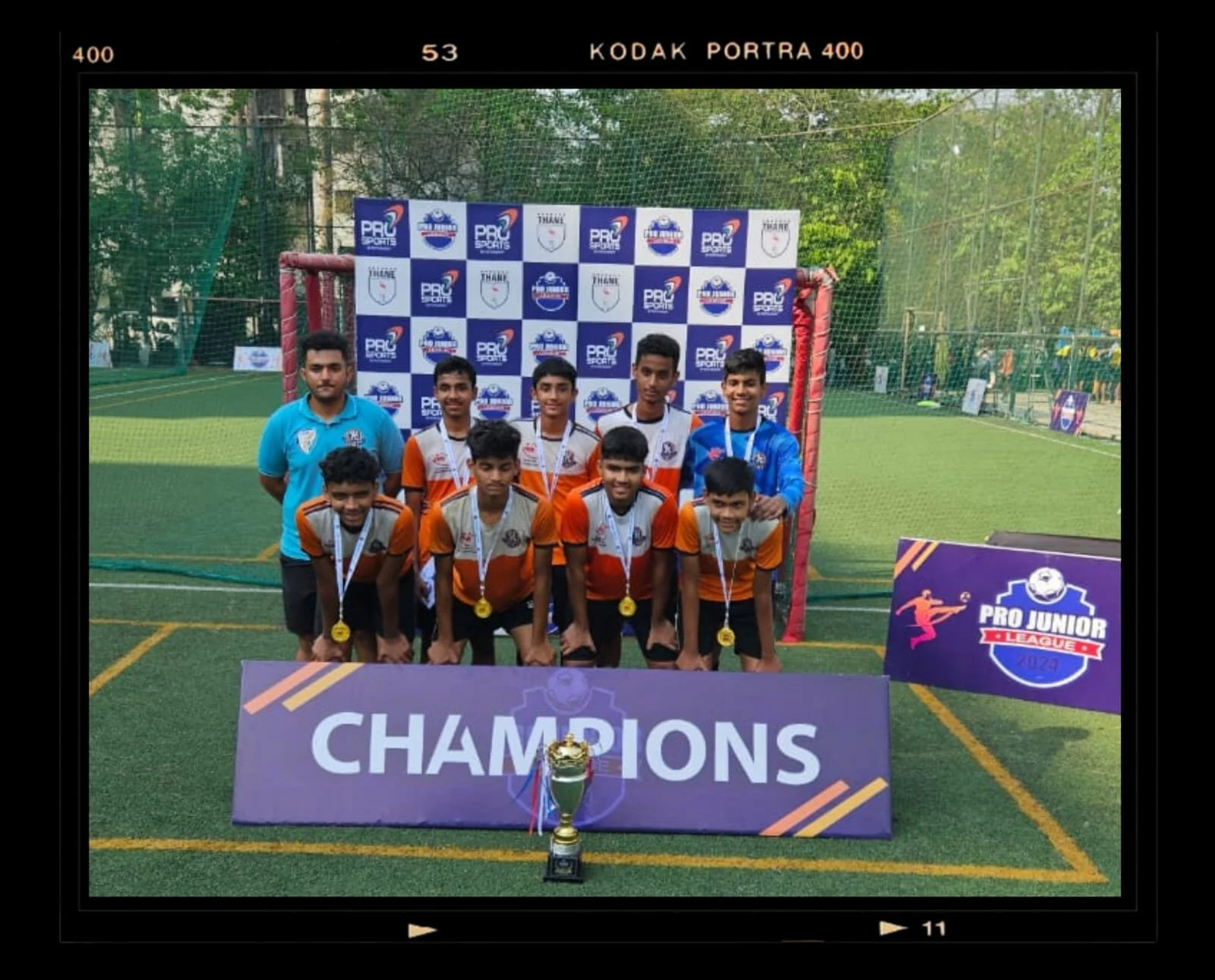 Youth soccer team posing with medals and a trophy on a field with a banner reading 'Champions' and 'Pro Junior League 2023' in the background.