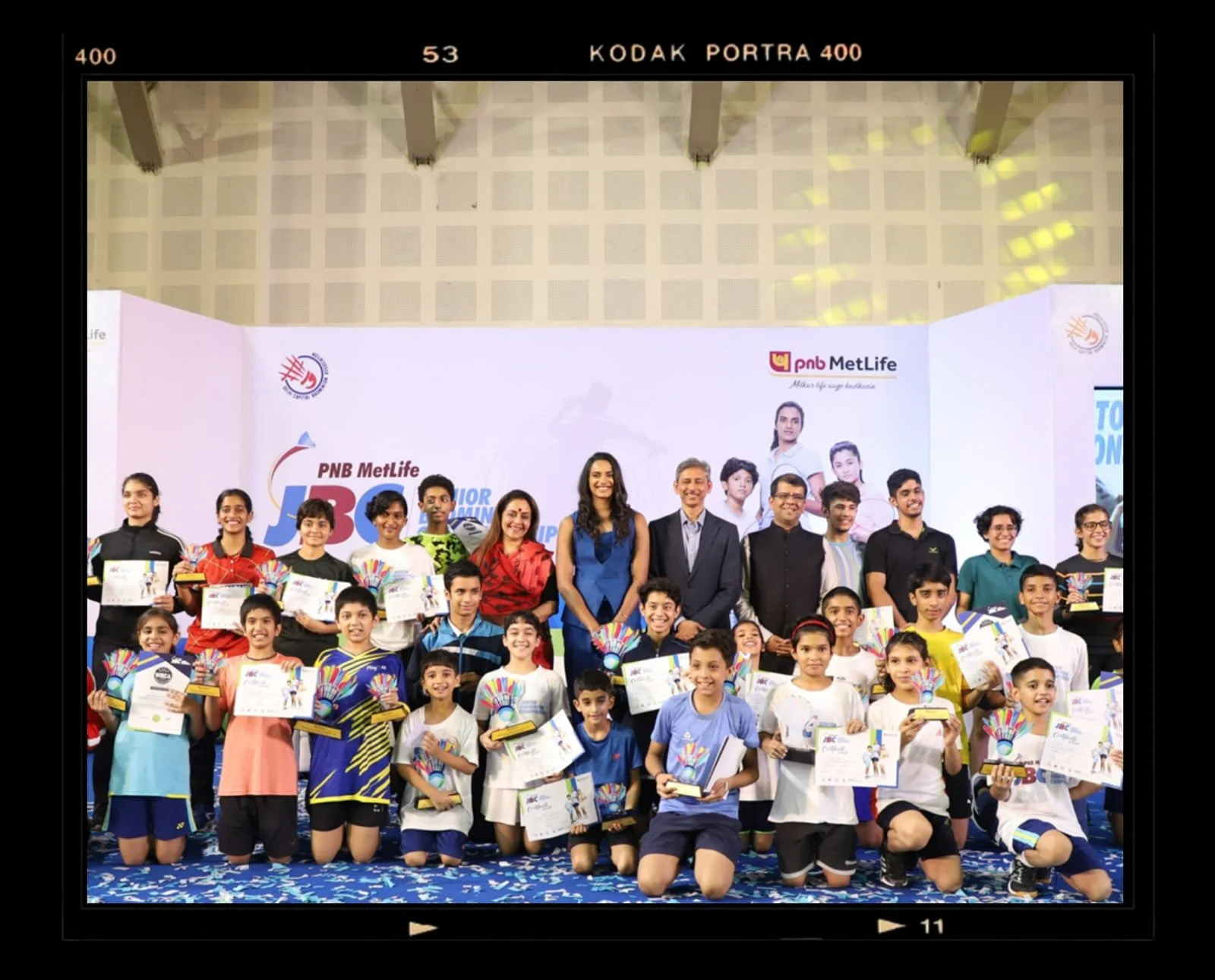 Group of children and adults posing with certificates and awards on stage at a children’s competition event, with a backdrop displaying event logos and sponsors.
