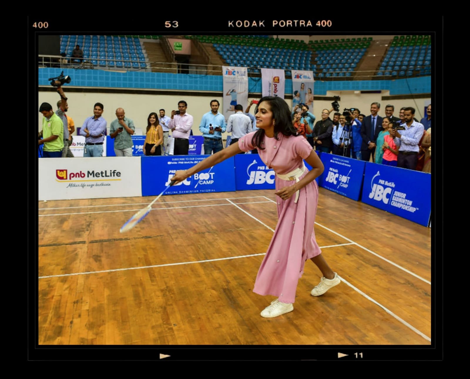 A woman in a pink dress and white sneakers stretching with a badminton racket on a wooden court during an indoor badminton event, with a crowd of people watching and photographers taking pictures in the background.
