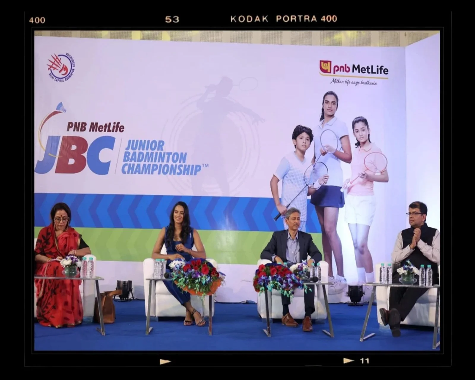 Panel at the PNB MetLife Junior Badminton Championship with five people sitting at a table, a large banner in the background displaying the event logo and images of children playing badminton, and a shadow of a person playing badminton on the banner.