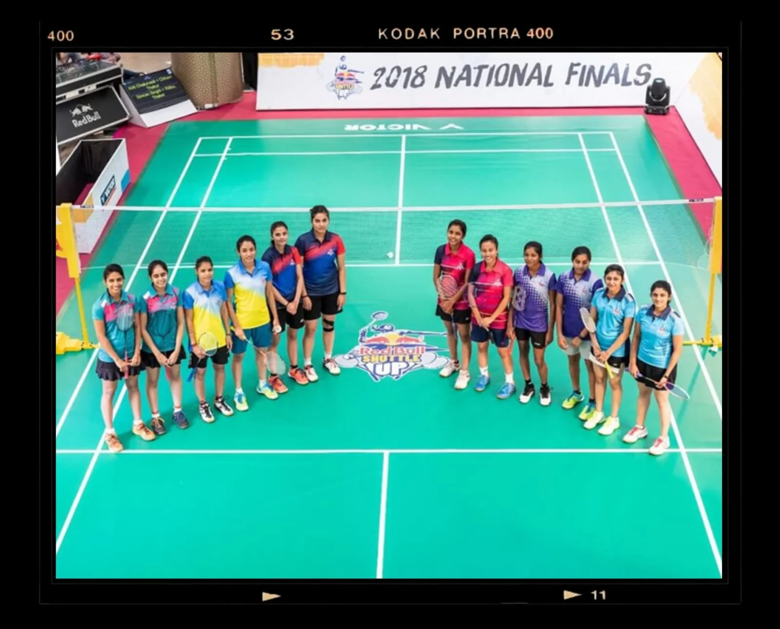 Group of young female badminton players standing in a line on a badminton court with a Red Bull logo and banner at the back, celebrating the 2018 National Finals of a badminton tournament.