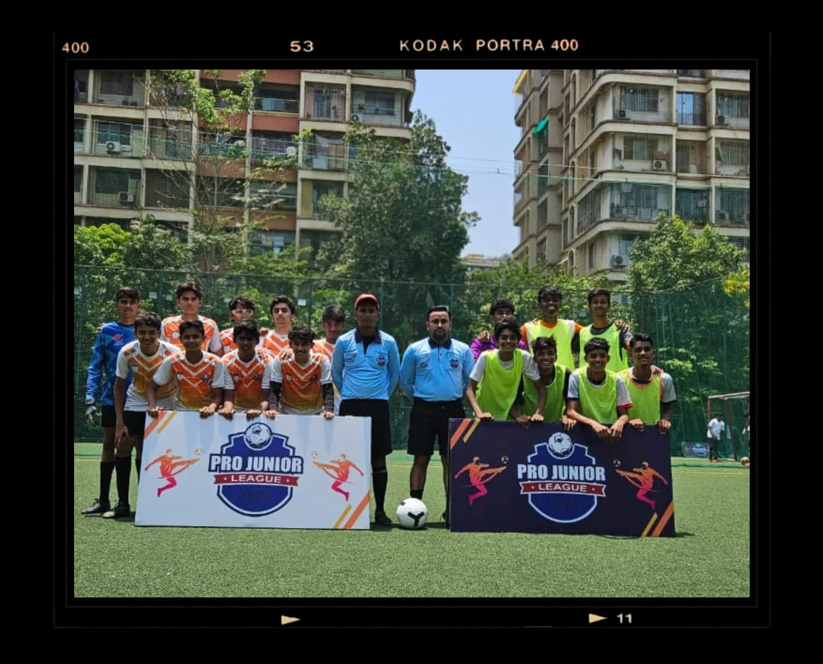 Group of young boys and two adult men standing on a soccer field, holding banners for 'Pro Junior League'. The boys are wearing sports jerseys and some with neon green bibs. The background shows tall apartment buildings and trees, with a blue sky.