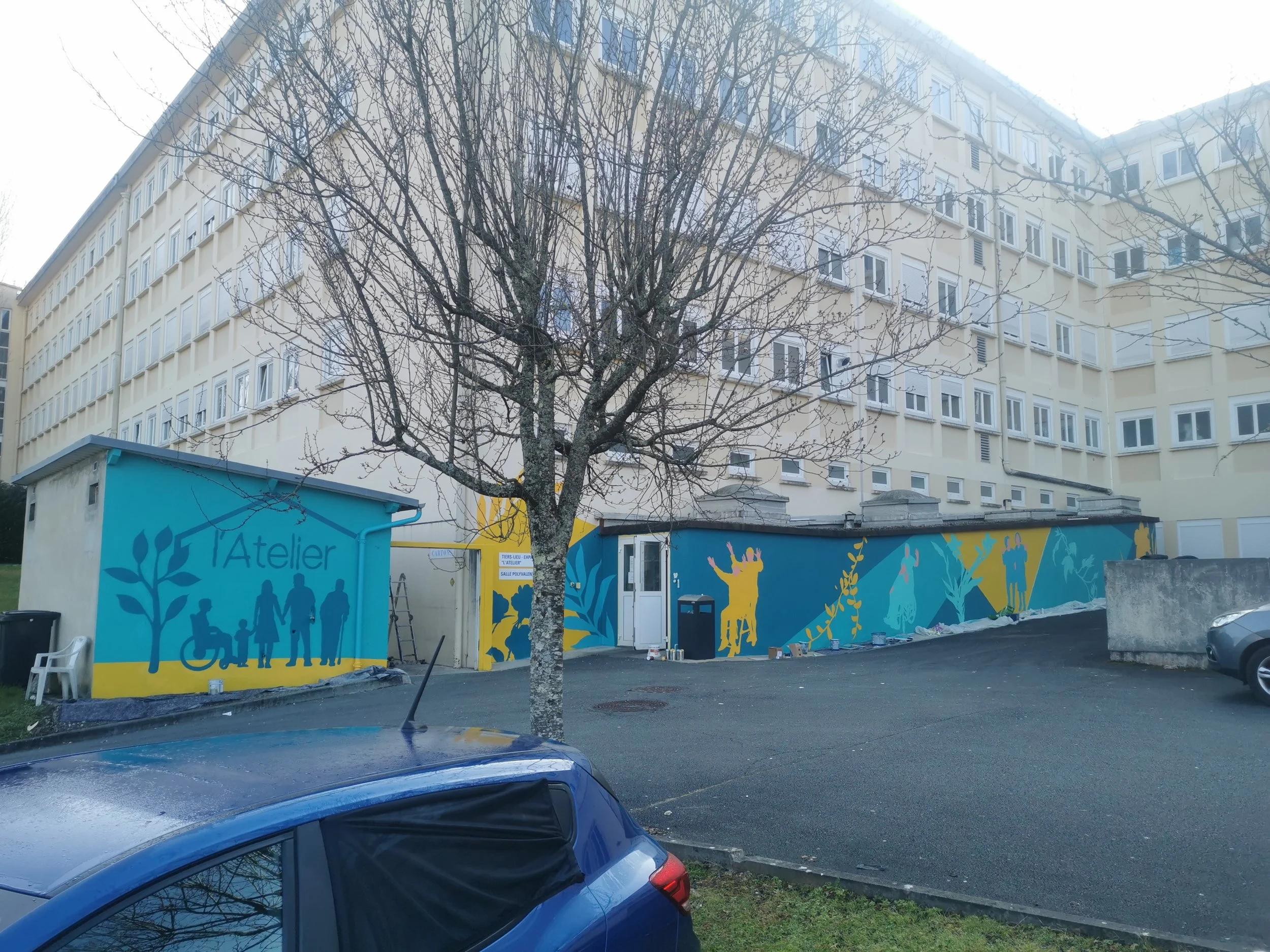 Vue d'un bâtiment résidentiel à plusieurs étages avec une école ou un centre communautaire en face, décoré avec des couleurs vives et des silhouettes humaines, et un arbre sans feuilles au centre. La scène inclut une voiture bleue au premier plan et des caisses de peinture posées près du bâtiment.