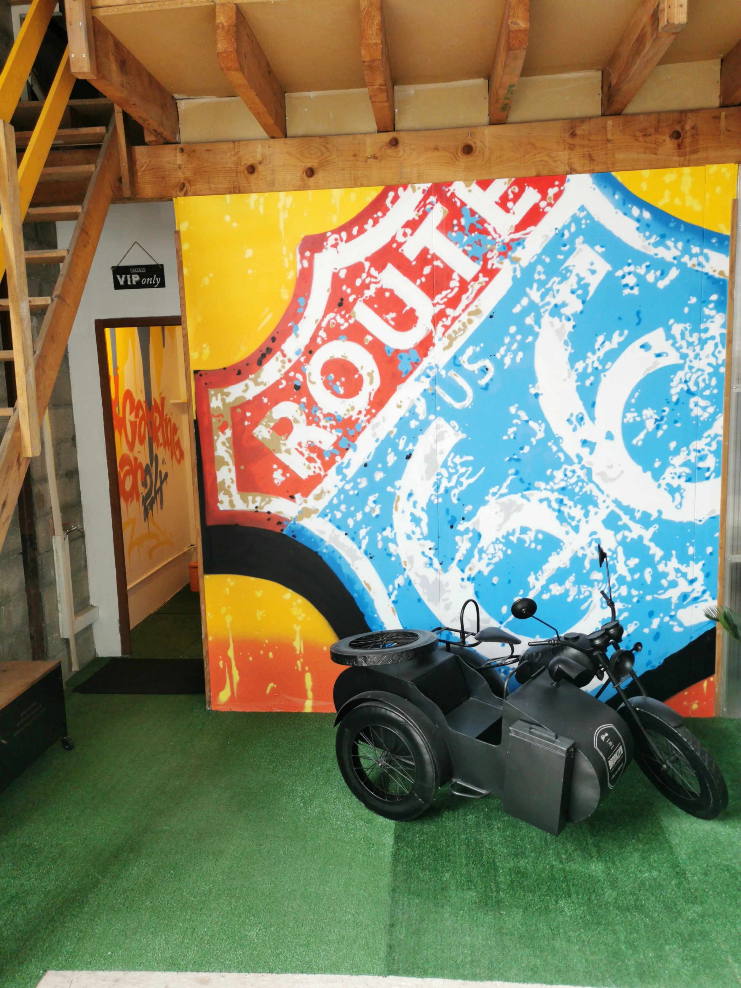 Vélo électrique noir avec un carénage gris, stationné sur un tapis vert artificiel devant un mur coloré avec du graffiti et un panneau « VIP only » en arrière-plan.