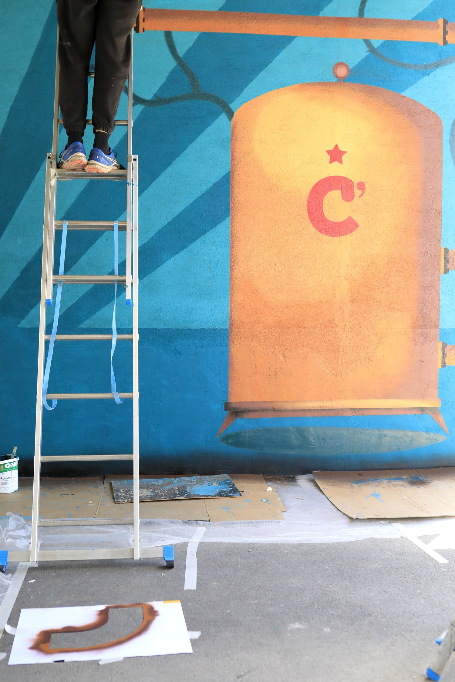 Un mur en train d'être peint avec un graffiti coloré représentant une grande  avec une étoile rouge et un symbole de C' au centre. Sur le sol, il y a des outils de peinture, un escabeau en aluminium avec une personne en haut et des feuilles de papier ou toiles avec des peintures ou esquisses.