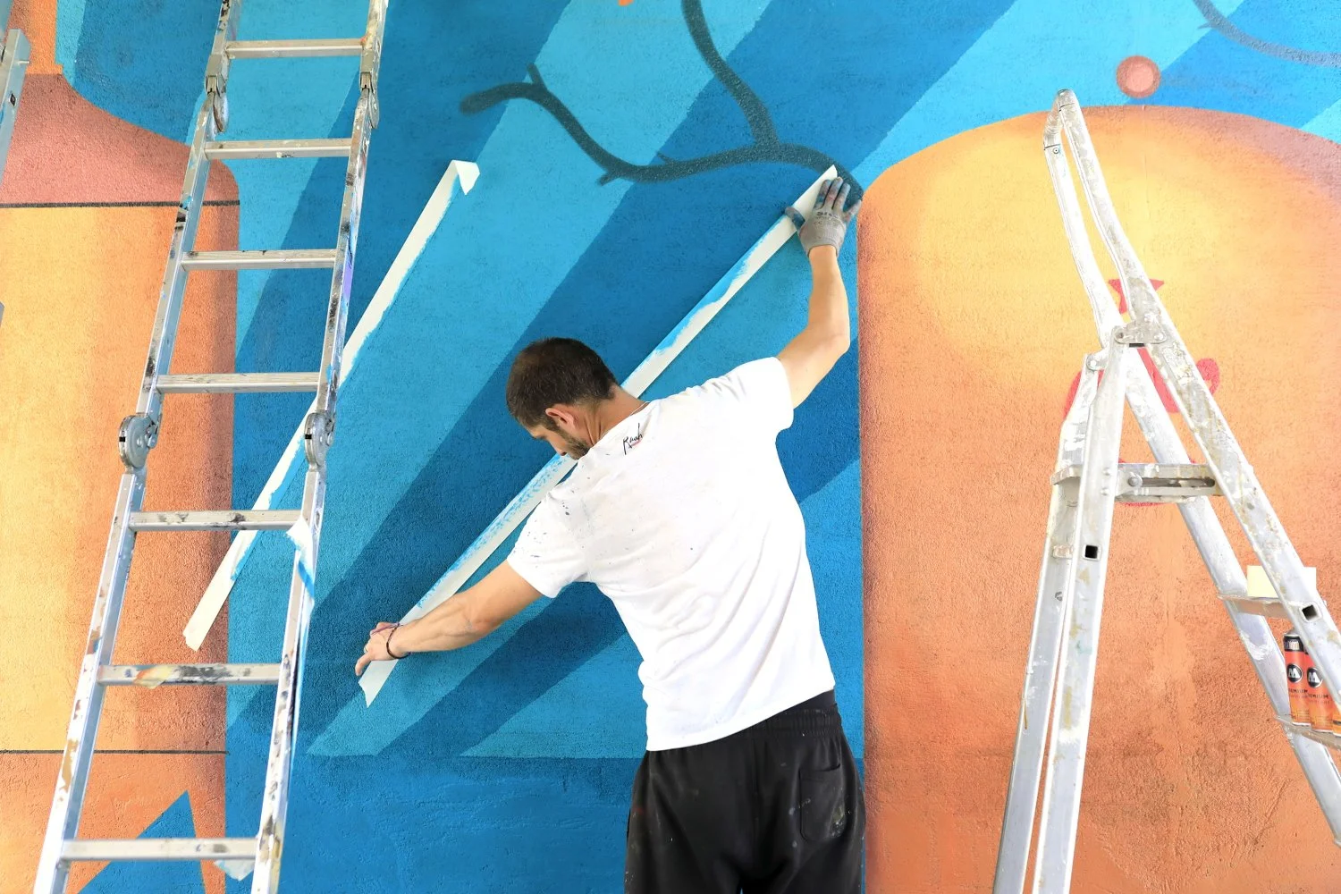 Un homme peint un mur coloré avec du bleu. Deux escabeaux en aluminium sont placés devant le mur, une à gauche et une à droite. Chavagn brasserie
