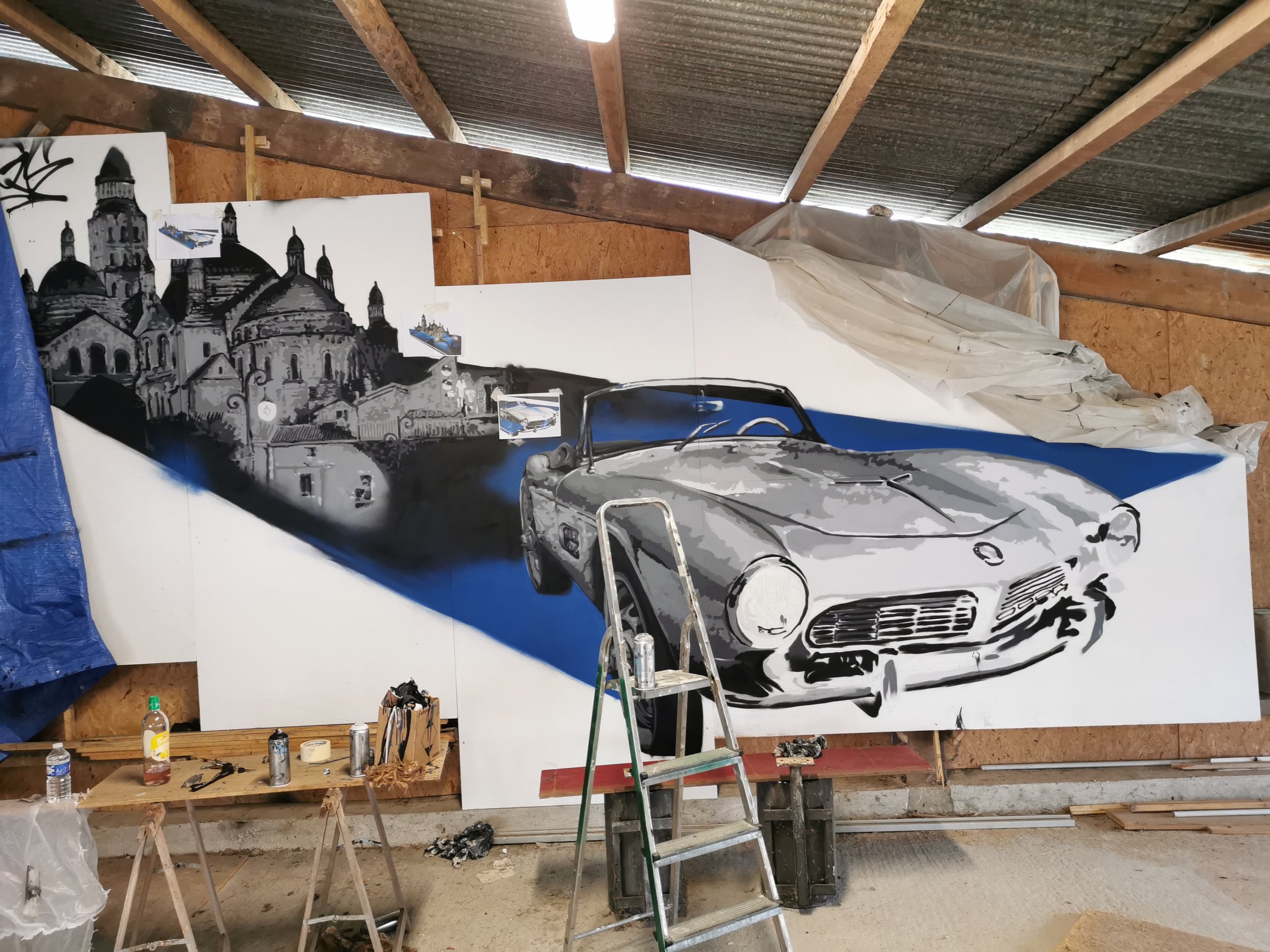 Une grande peinture représentant une voiture de sport devant une vue d'une ville historique avec un bâtiment religieux. La scène est en noir et blanc avec des touches de bleu. La peinture est en cours de réalisation dans un atelier avec des outils et des bouteilles d'eau sur un tapis de sol, et un escabeau.