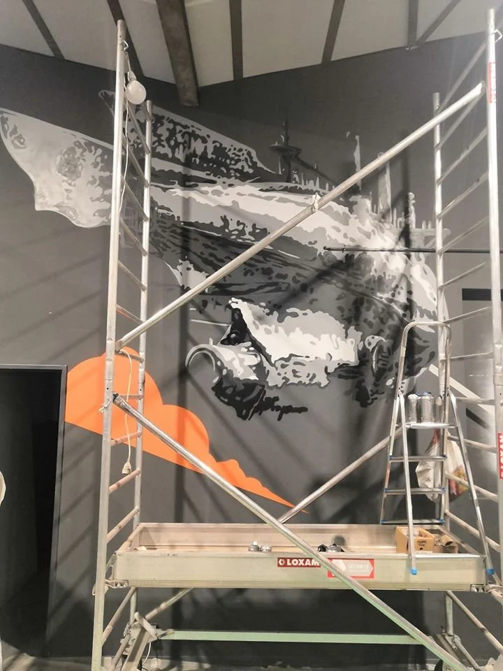 Mur en cours de peinture représentant un visage en noir et blanc avec une explosion orange au bas, échafaudage à côté.