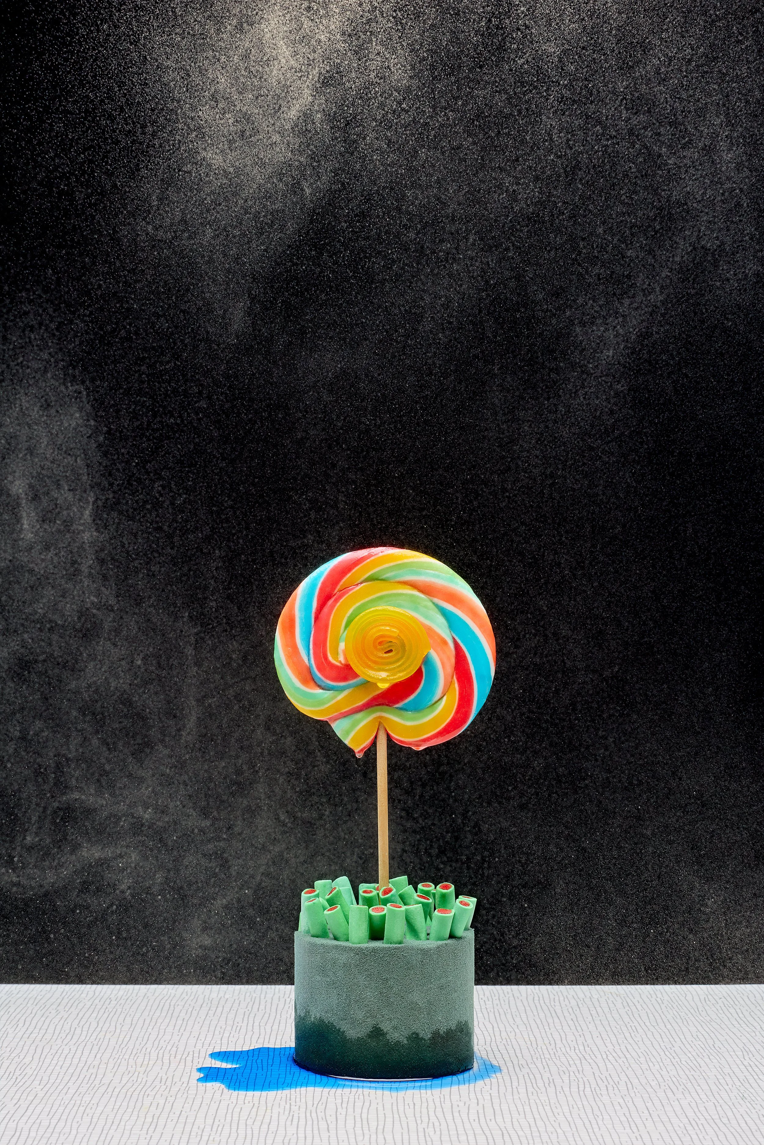 Colorful swirled lollipop on a stick inside a container filled with green sweets, against a black starry background.
