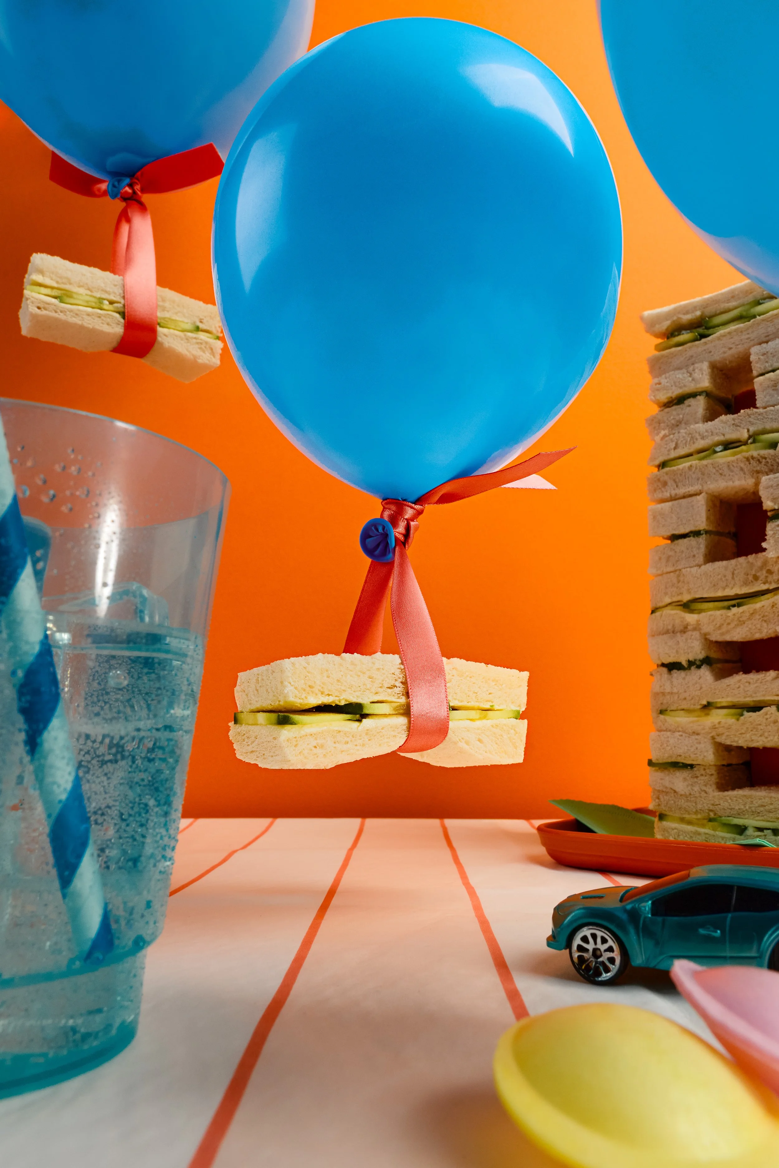 Colorful birthday party scene with blue balloons tied with ribbons, small sandwiches, a glass of water, a toy car, and a yellow balloon on a table with orange and white background.