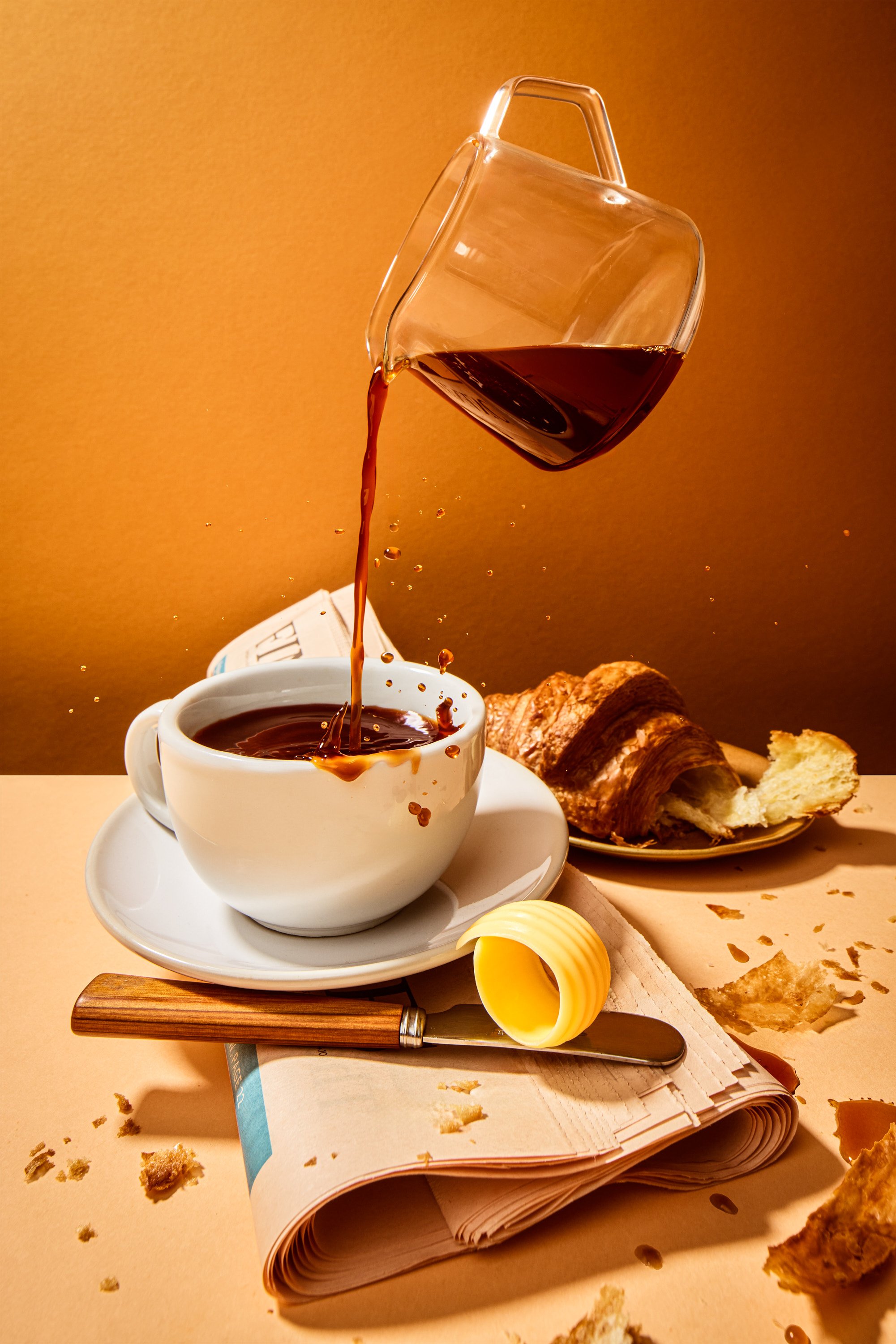 A cup of black coffee with a book, a croissant, and a croissant roll with crumbs scattered on a light-colored table. A glass of dark liquid is being poured into the cup from above, and there is a yellow butter curl near the book.