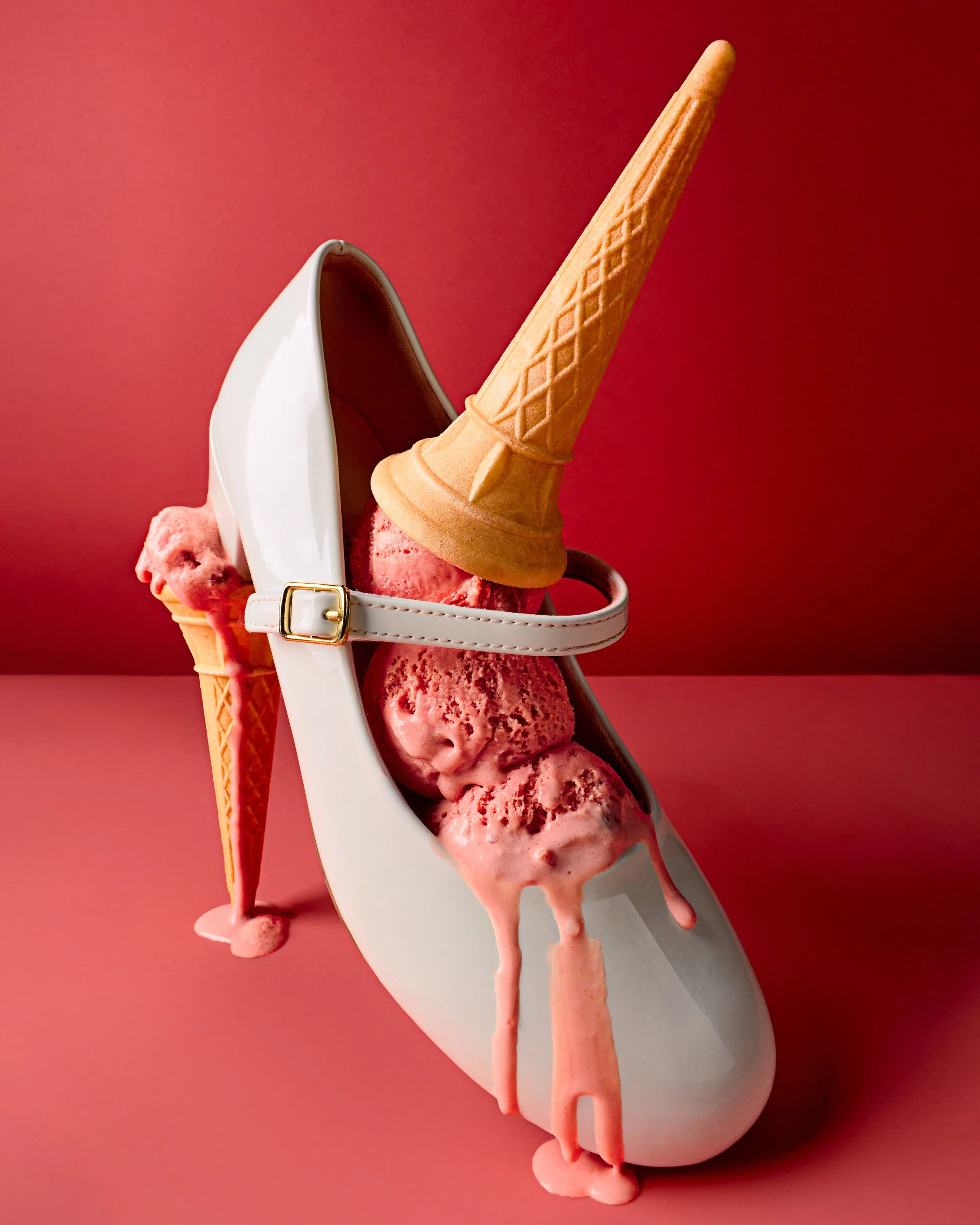 A white child's shoe filled with three scoops of pink ice cream. An ice cream cone is placed upside down on the ice cream and as the heel of the shoe, and pink ice cream drips down from the scoops onto a pink surface, with a solid pink background.