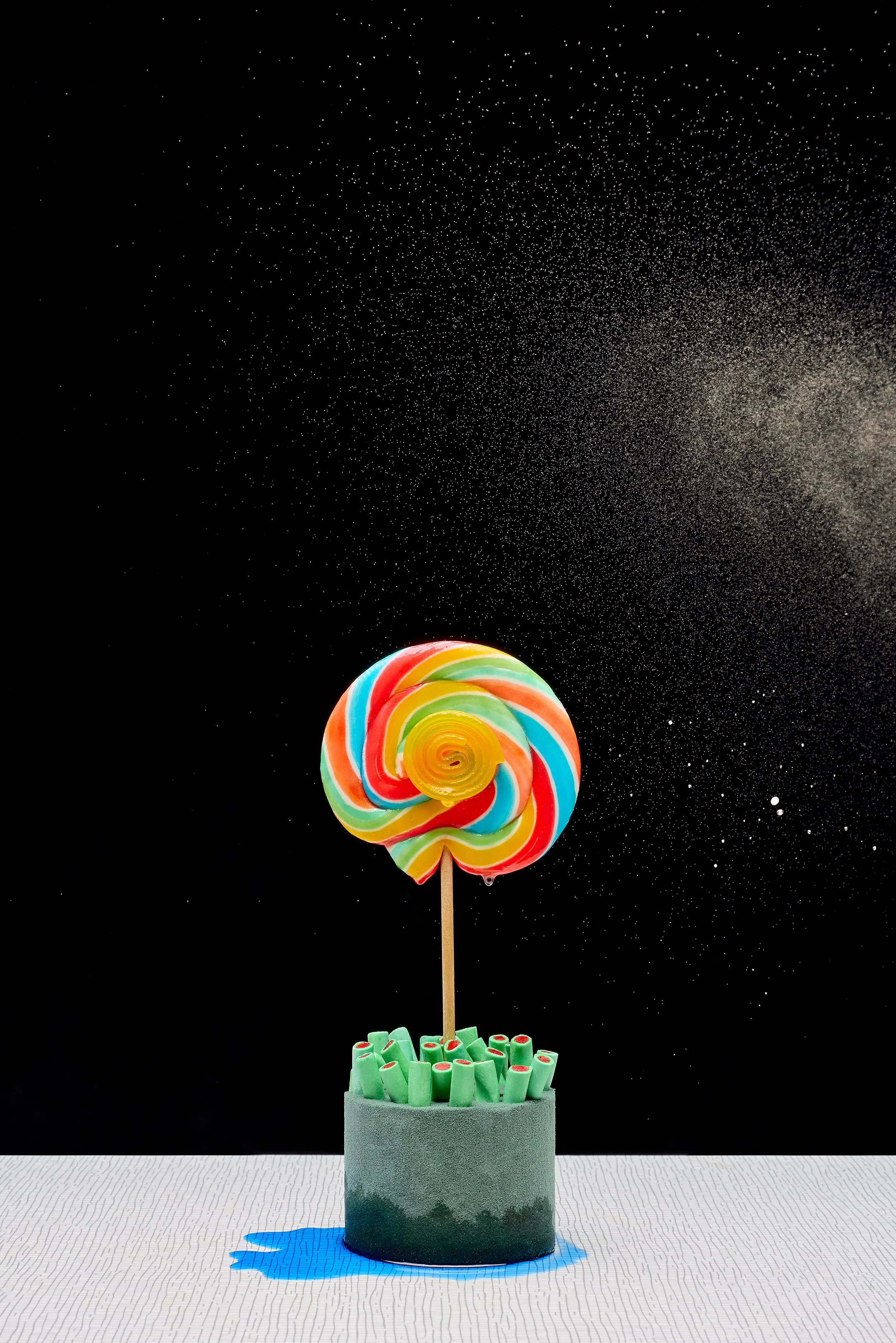 A colorful rainbow swirl lollipop on a stick rising from a gray container filled with green candy rolls, set against a black background with a scattering of white specks resembling stars, and a blue shadow beneath the container.