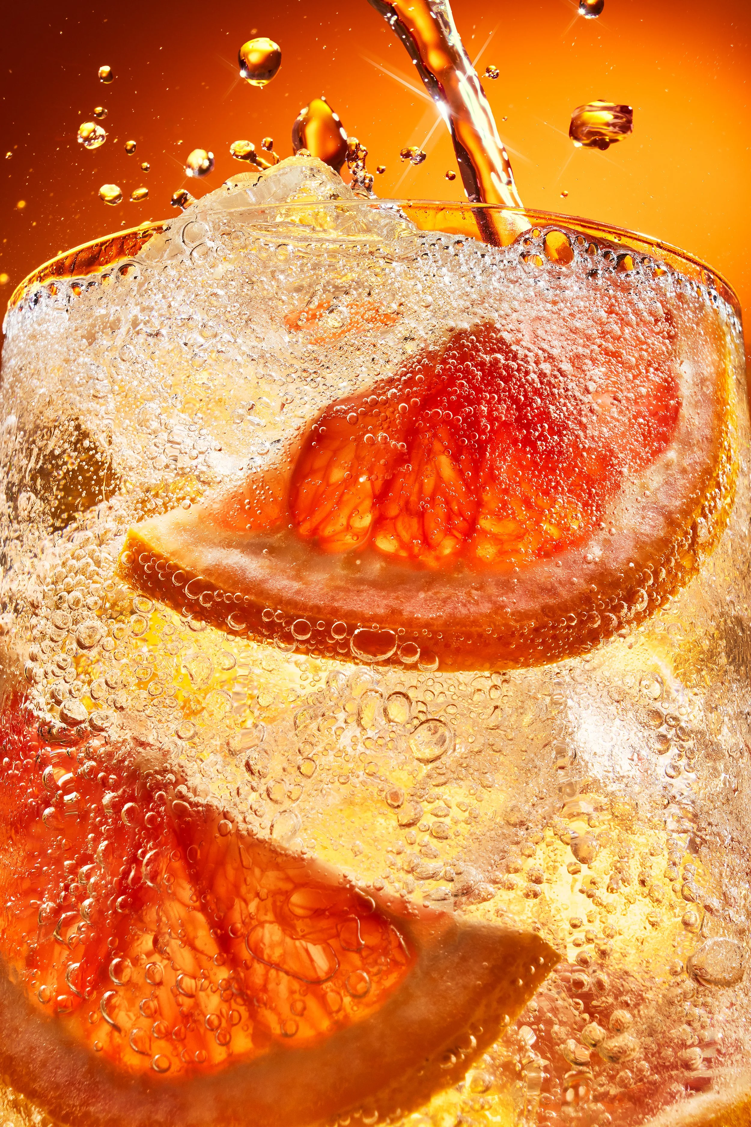 Close-up of a glass with ice, orange slices, and a dark soda being poured into it, creating bubbles and a glowing amber background.