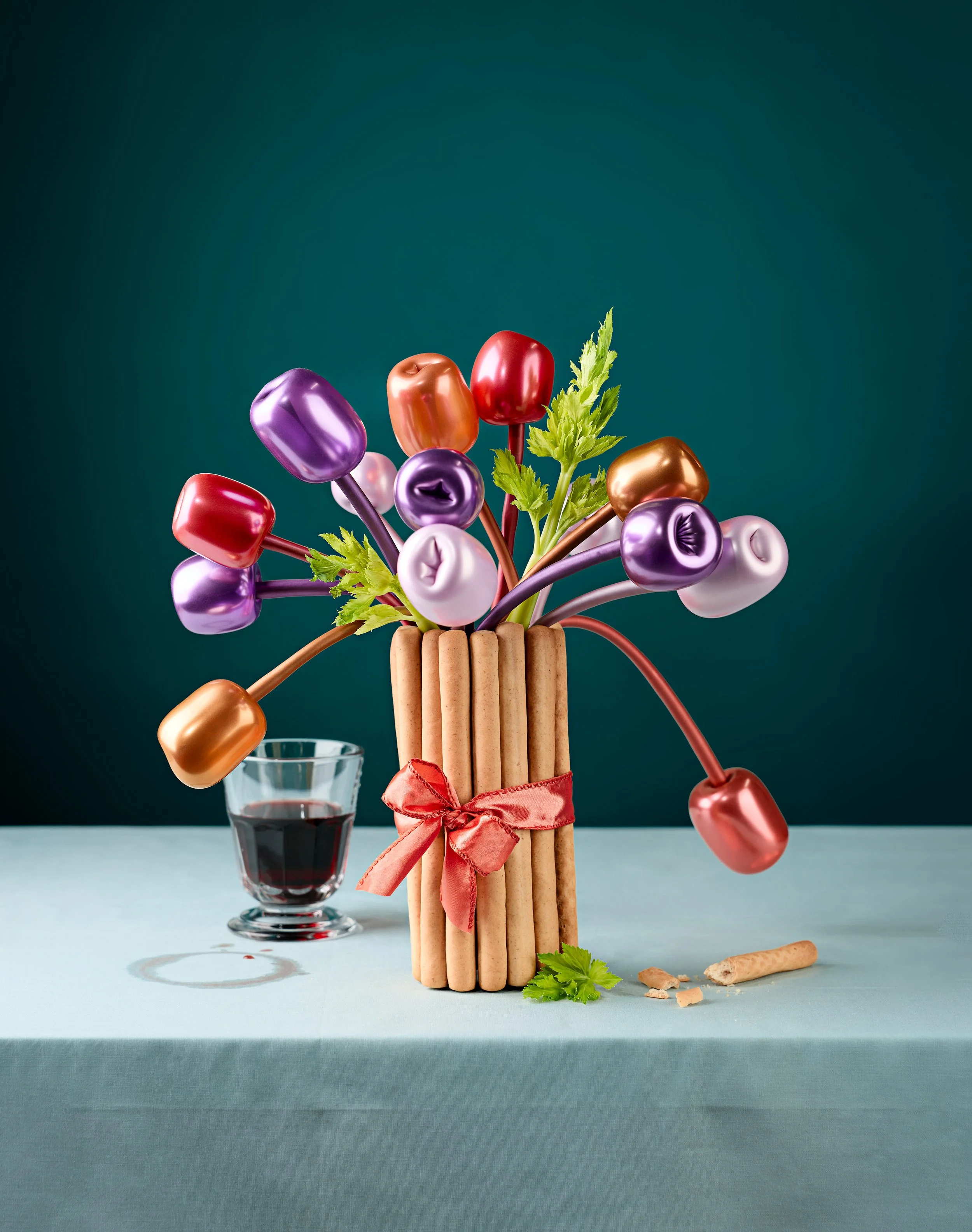 Arrangement of balloon tulips and celery in a vase made of bread sticks, with a glass of red wine and scattered breadsticks and a sprig of celery on a light blue surface against a dark teal background.