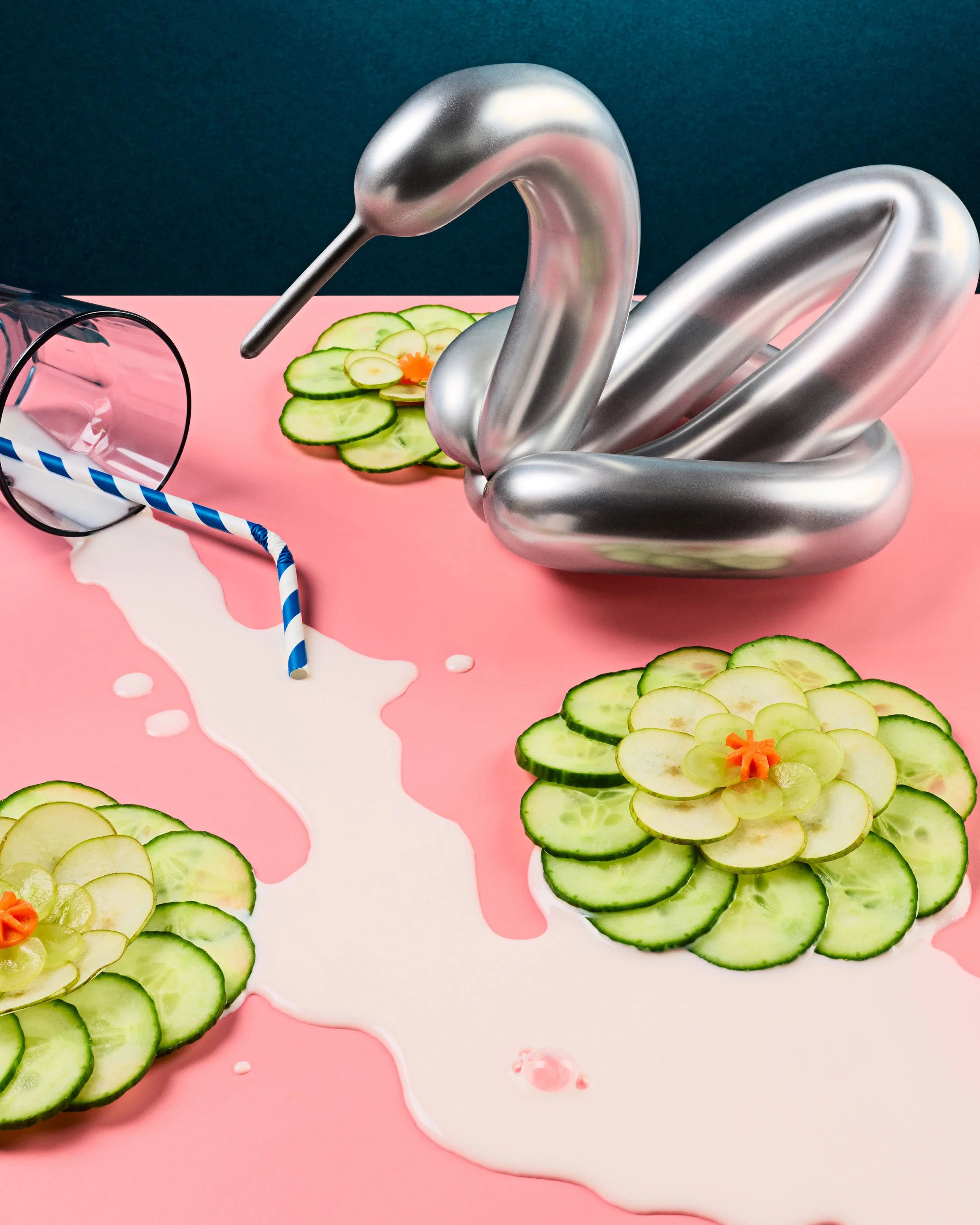 A close-up image of a silver swan-shaped balloon on pink surface with cucumber slices and carrot flower, with spilled milk and blue-and-white striped straw.