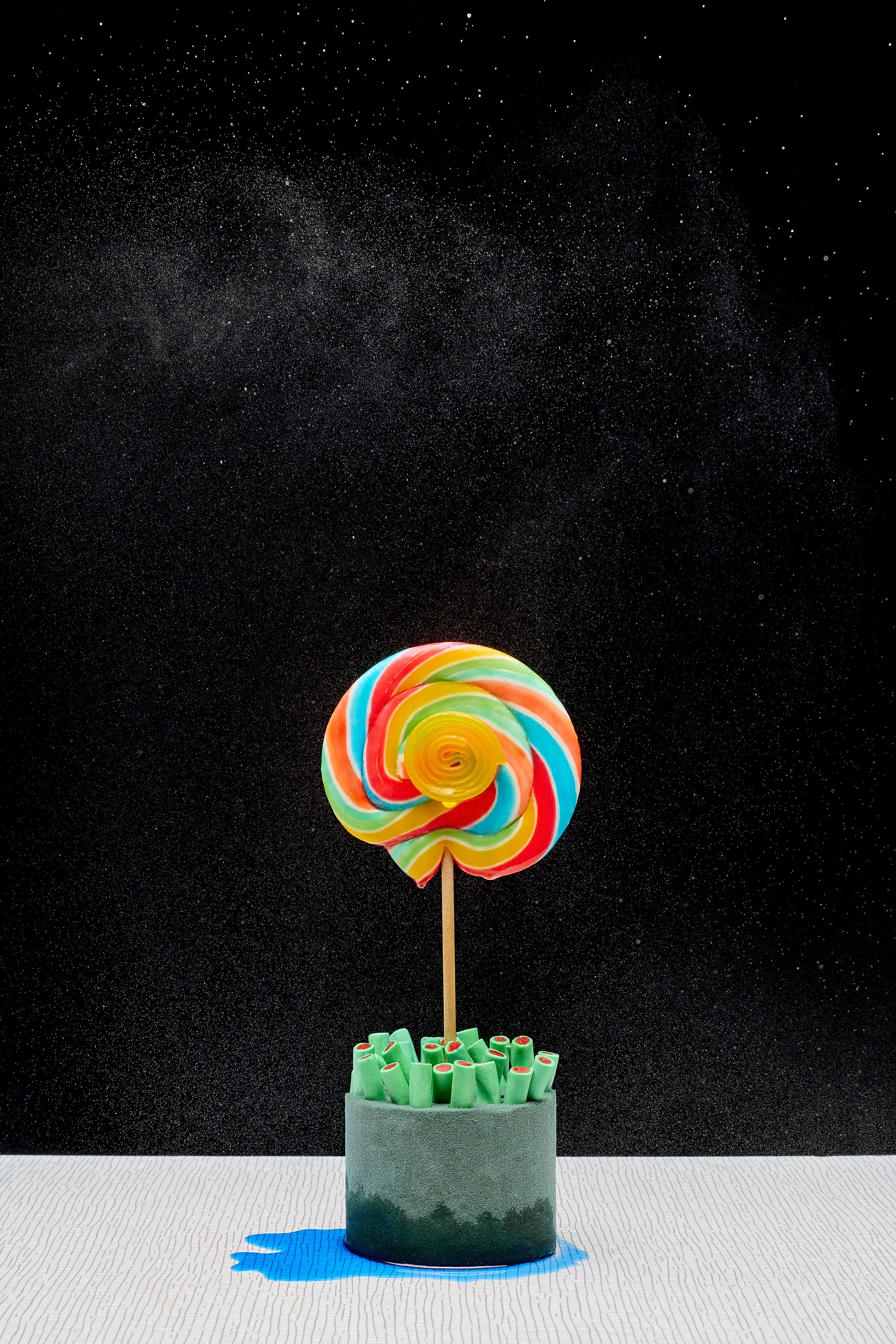 Colorful swirled lollipop on a stick inside a container filled with green sweets, against a black starry background.