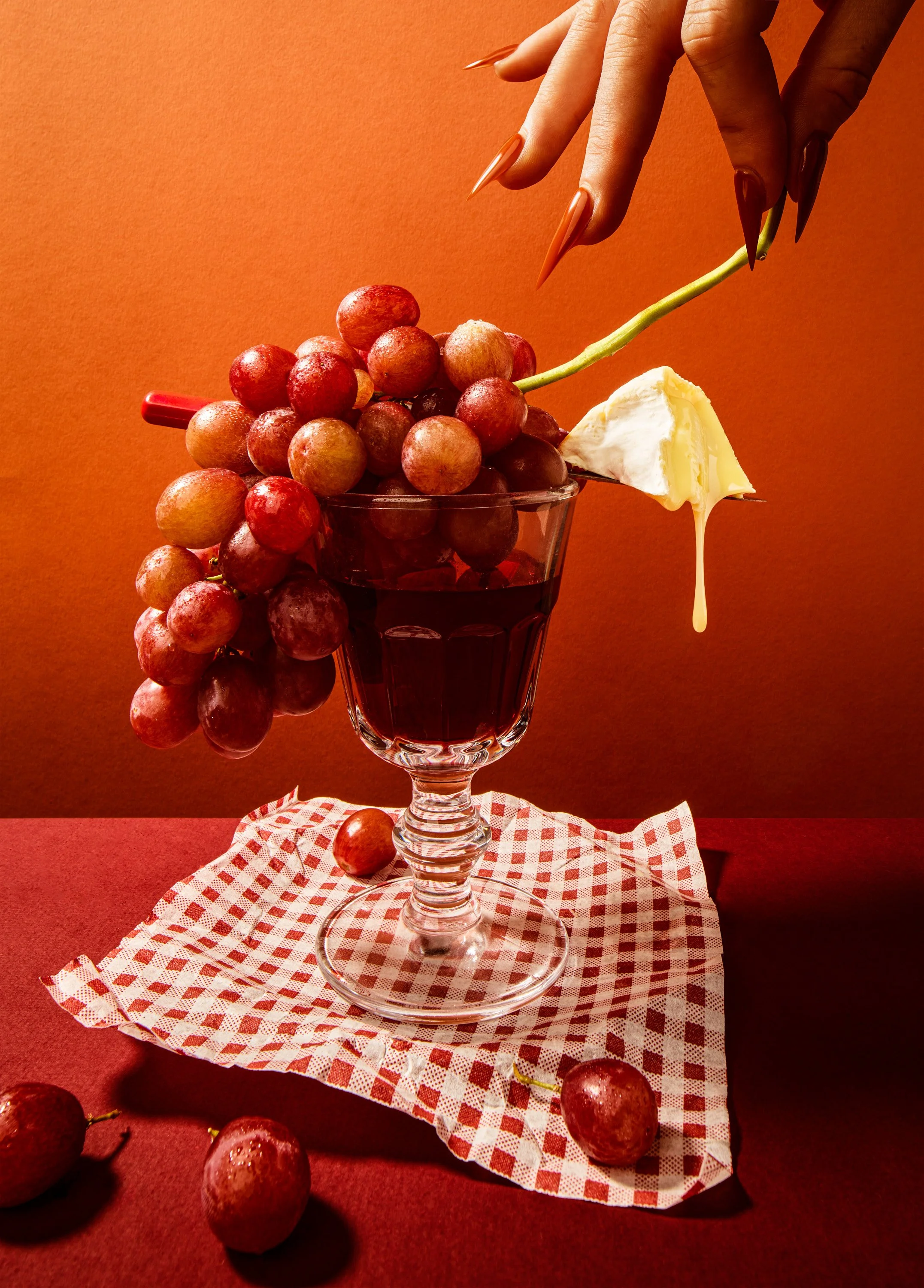 A glass of red wine with red grapes, a wedge of Brie cheese, and a hand with long, sharp, brown nails reaching towards the grapes and cheese on a red and white checkered napkin.