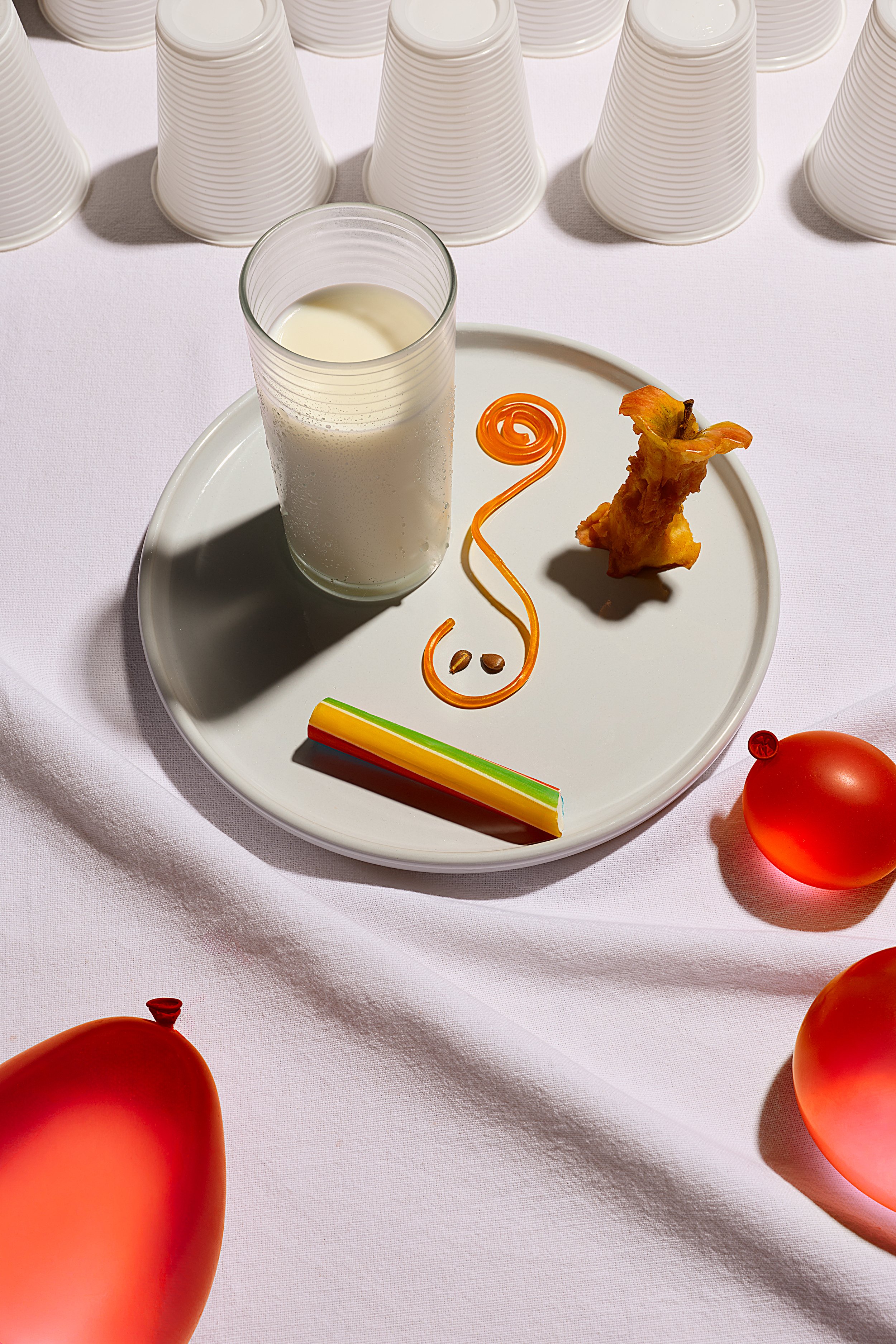 A white plate with a glass of milk, an eaten apple, a swirl of orange sweet lace and a colorful striped rock candy on a white tablecloth. Several red balloons are visible around the plate.