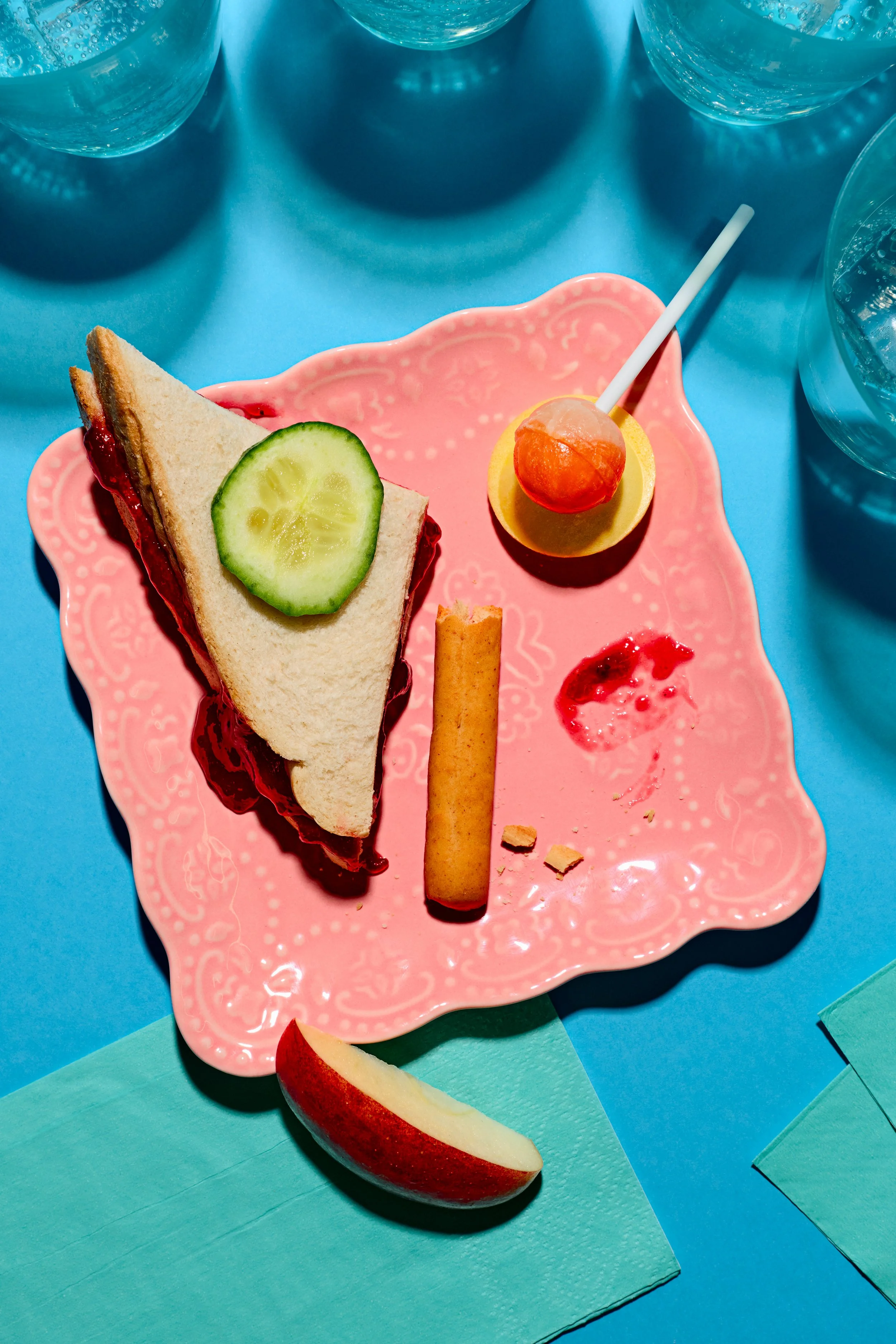 A pink decorative plate holding a sandwich with cucumber and jam, a popcicle, and a breadstick with crumbs, on a blue surface with blue water glasses around. 