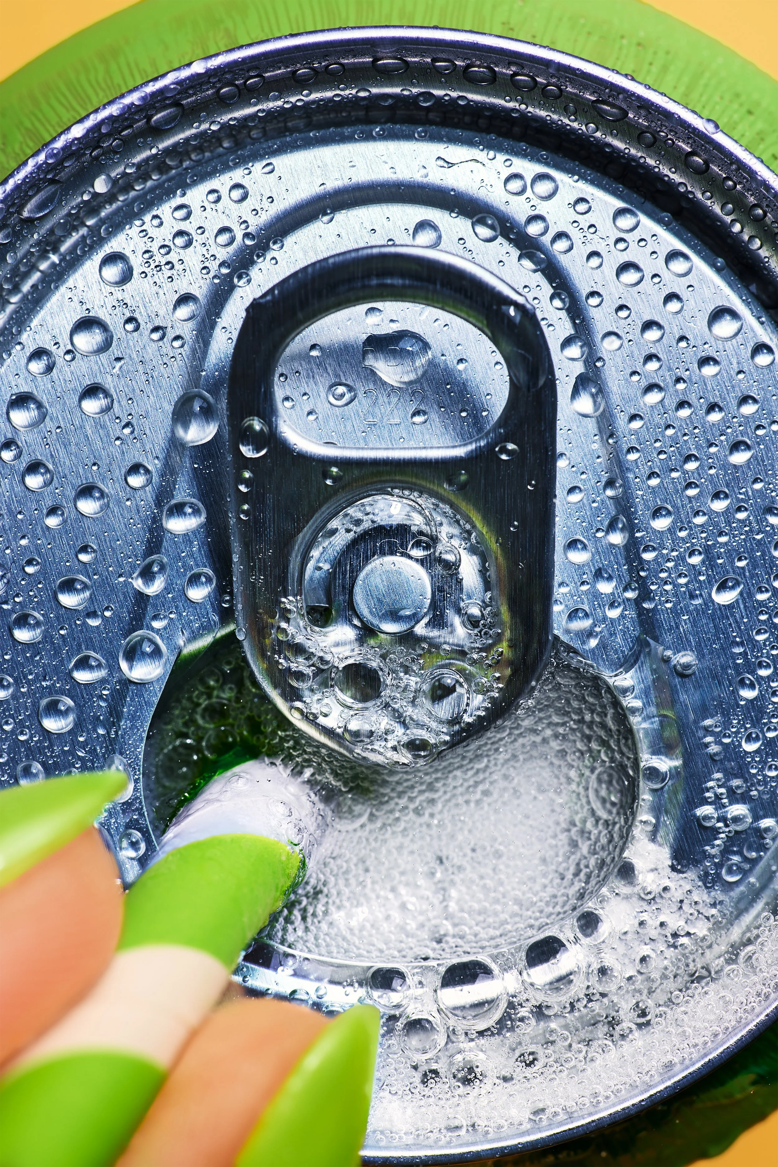 Close-up of the top of a soda can with condensation and drops of water on the surface, and a person's fingers with green painted nails holding the can.