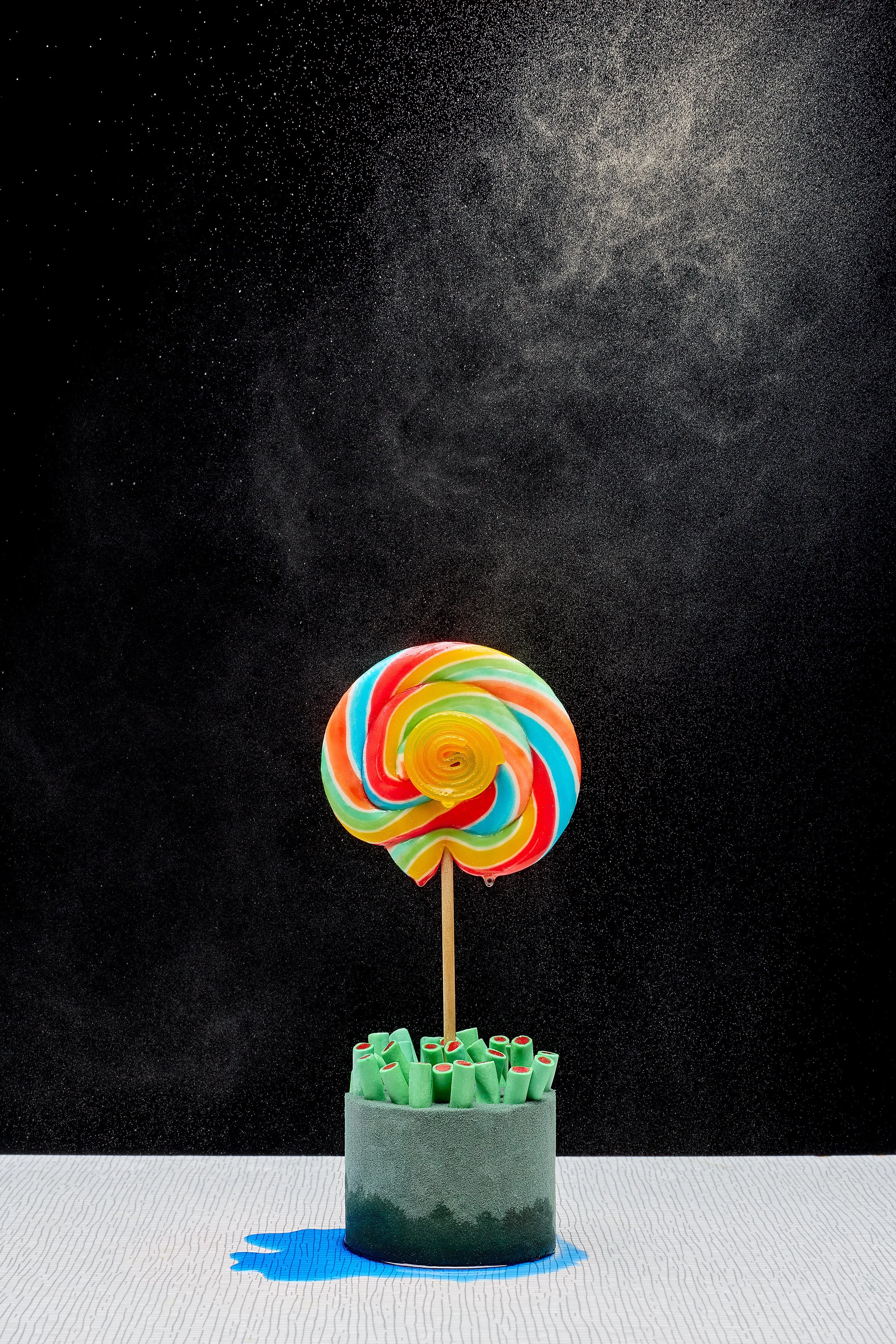 Colorful swirled lollipop on a stick inside a container filled with green sweets, against a black starry background.