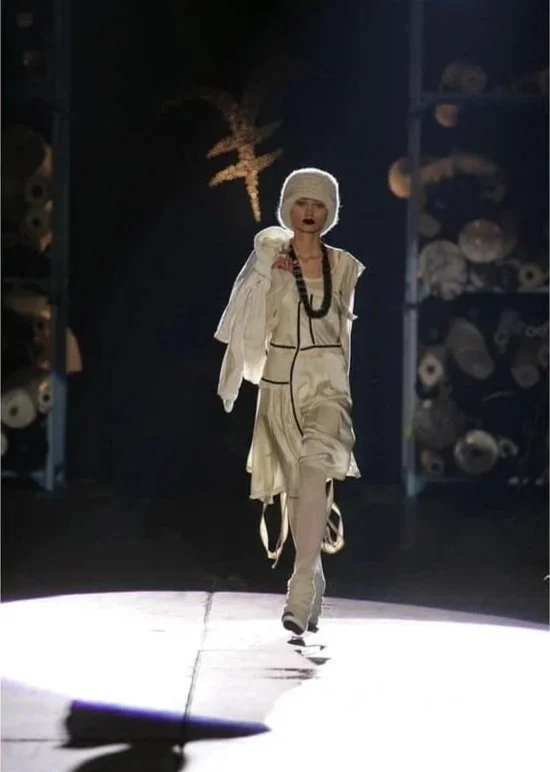 Modella che cammina sulla passerella, indossando abbigliamento bianco con cappello e accessori, in uno scenario artistico con luci e decorazioni sullo sfondo.