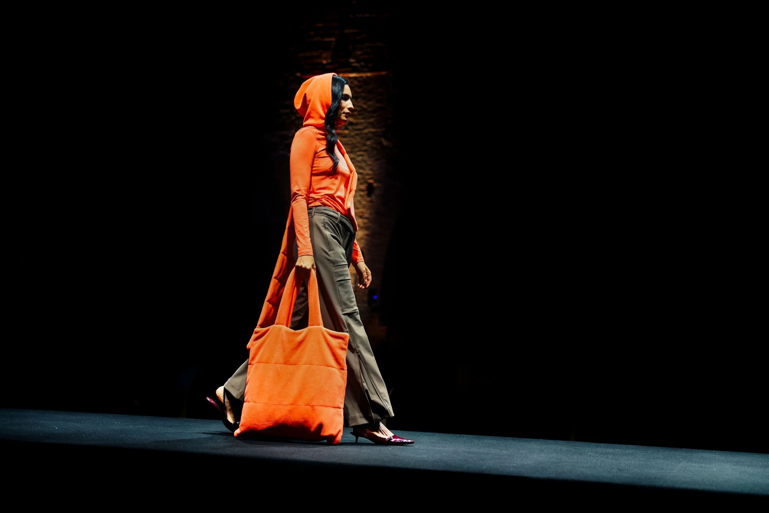 Una donna in passerella indossa pantaloni cachi con stivali, una mantella e borsa arancione, con alle spalle una lampada e uno sfondo scuro.