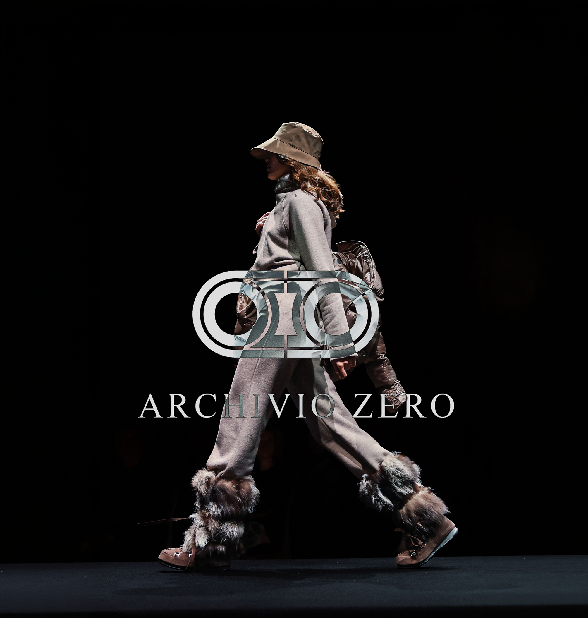 Modella in passerella con cappello beige, giacca grigia, pantaloni beige larghi con pelliccia alle scarpe, sfondo nero.