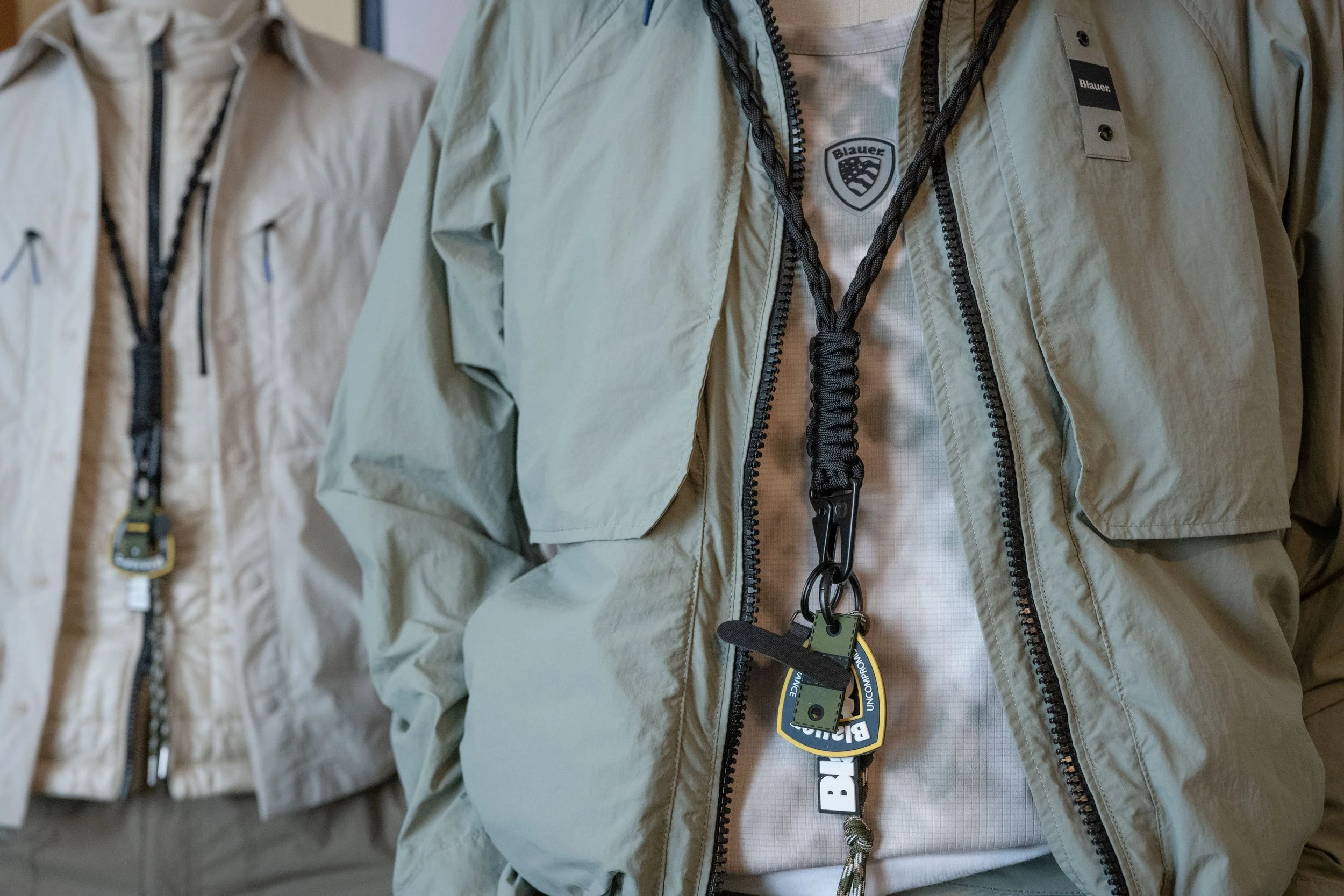 Immagine di diverse giacche color beige appese in una fila, con una in evidenza al centro che mostra un'etichetta e due tag di brand. Le giacche hanno tasche con zip e dettagli come cordini e utili ganci porta oggetti.
