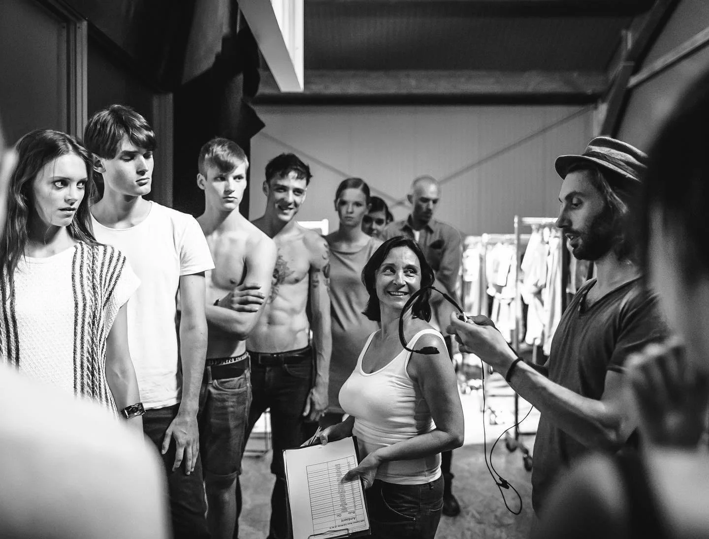 Un gruppo di persone in una sala di scena o studio di moda, alcune in abiti casual e altre senza maglietta, ascoltano con attenzione mentre una donna e un uomo discutono e ridono, con quest'ultimo che tiene in mano un dispositivo elettronico.