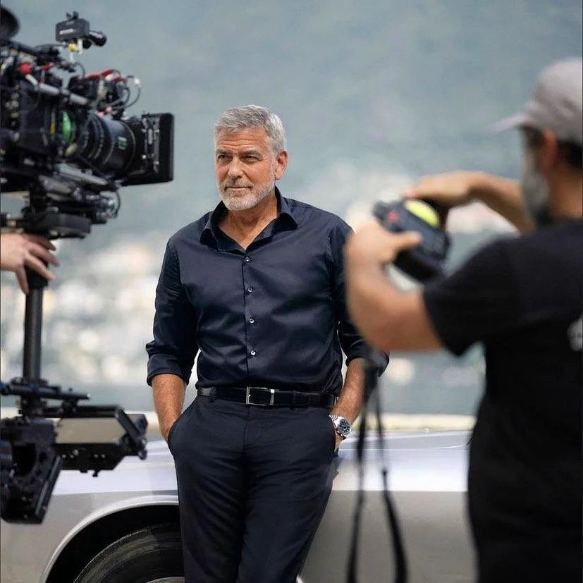 George Clooney vestito in camicia nera, durante un'intervista o una ripresa video all'aperto, con due persone che lo stanno filmando. Sfondo sfocato di montagne o collina.