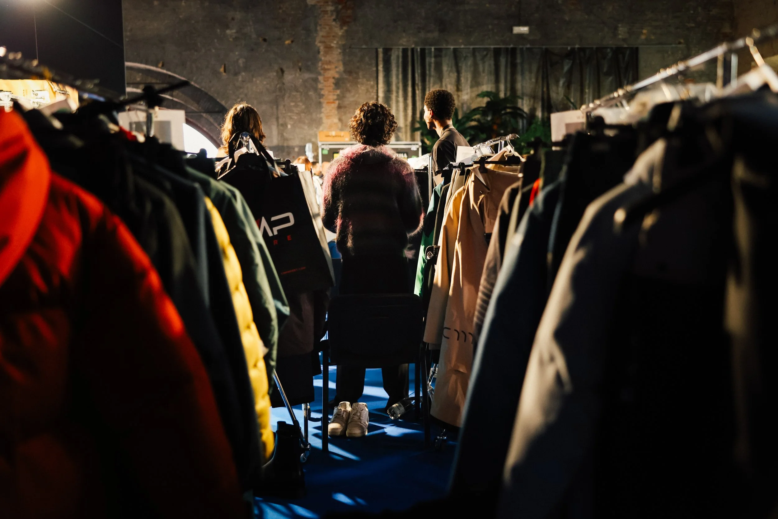Persone in fila tra scaffali di abbigliamento, vista da dietro, in un ambiente con pareti di mattoni e illuminazione calda.